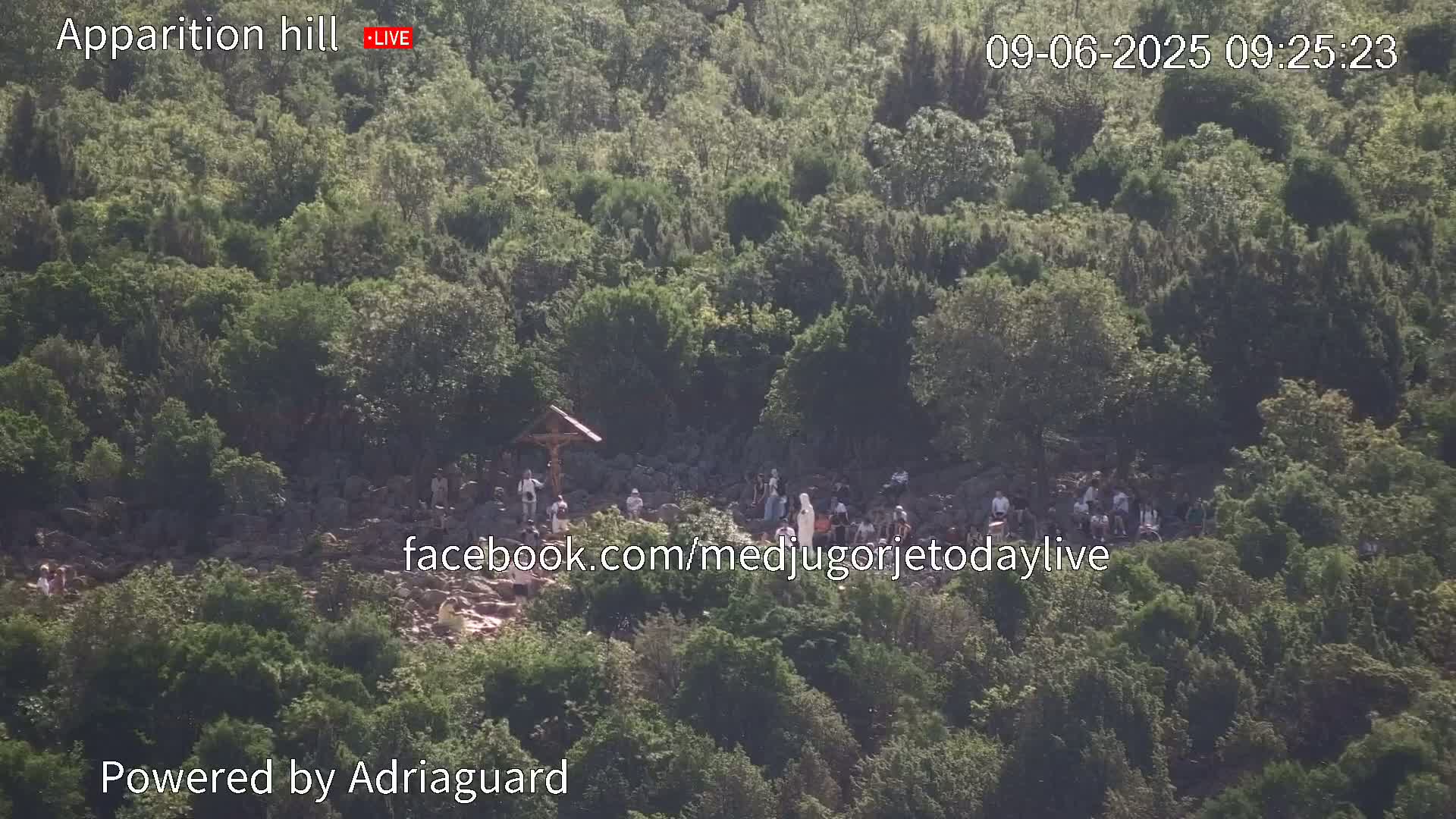 A hillside covered in lush green vegetation shows a group of people gathered near a crucifix under a sunny sky.