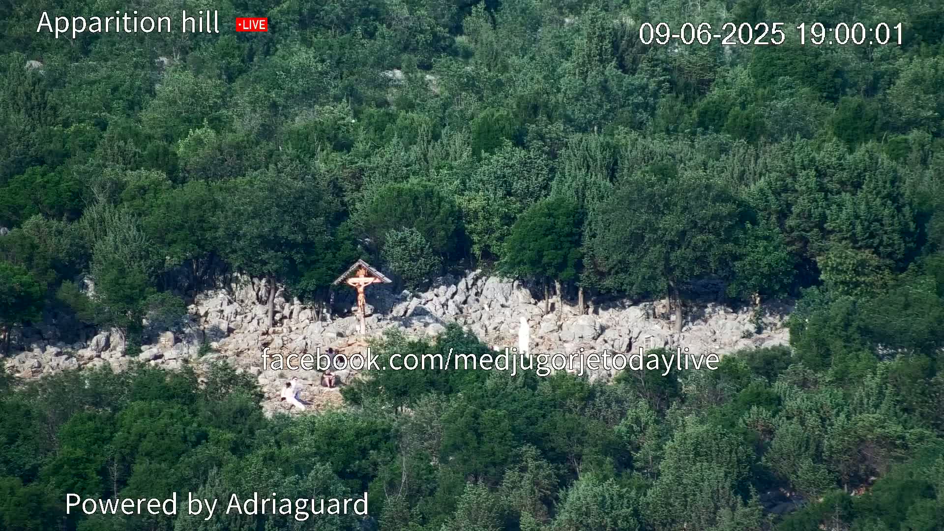 A rocky hillside covered in lush green trees features a crucifix and a statue, with several people visible near the base.
