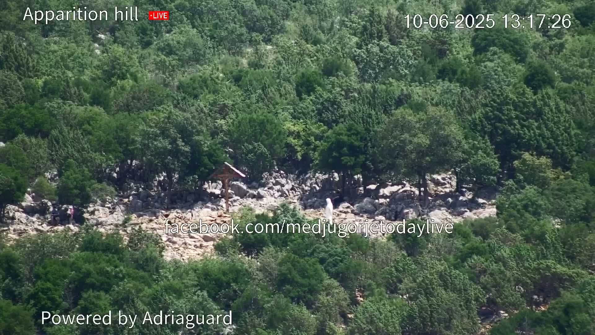 A rocky hillside covered in lush green trees and shrubs, with a few people visible on a path, features a wooden cross and a white statue.