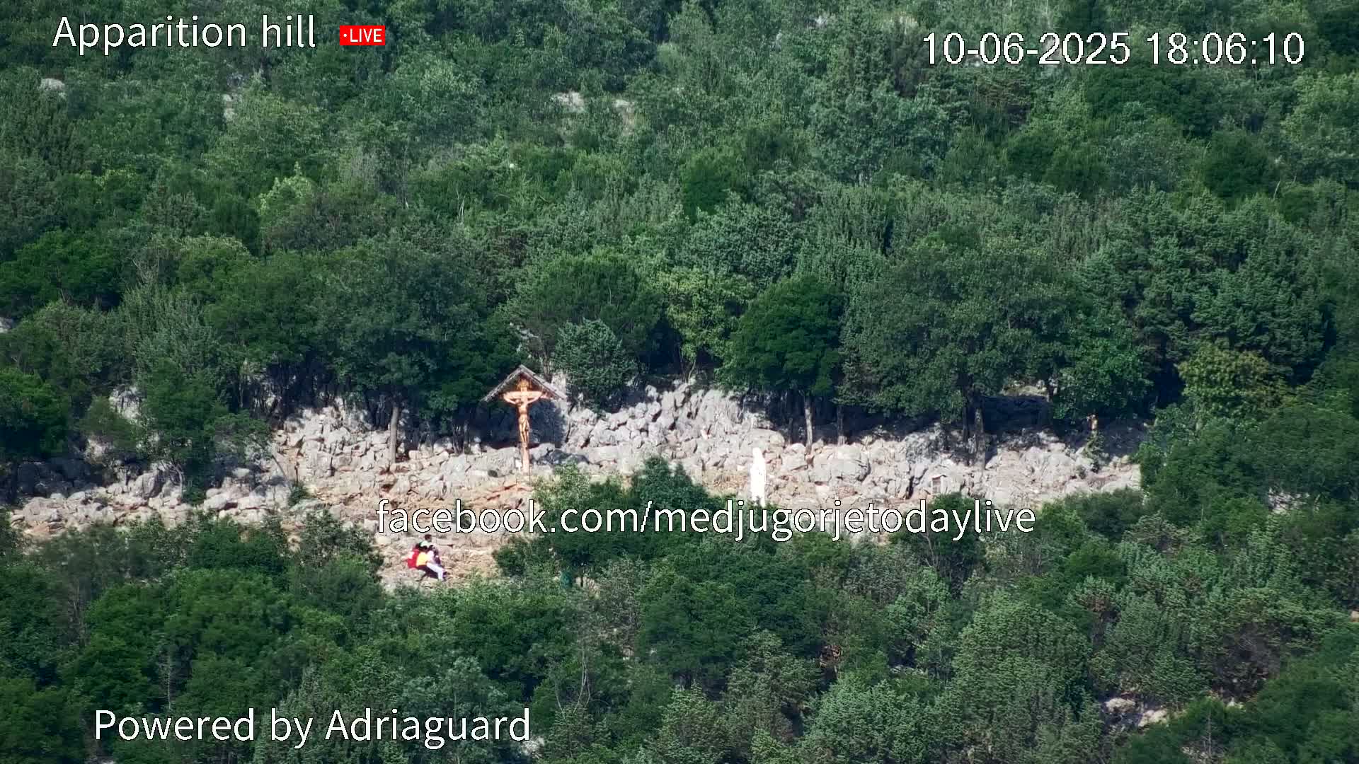 A rocky hillside covered in green trees and shrubs features a wooden crucifix and a white statue, with a few people visible near the base.