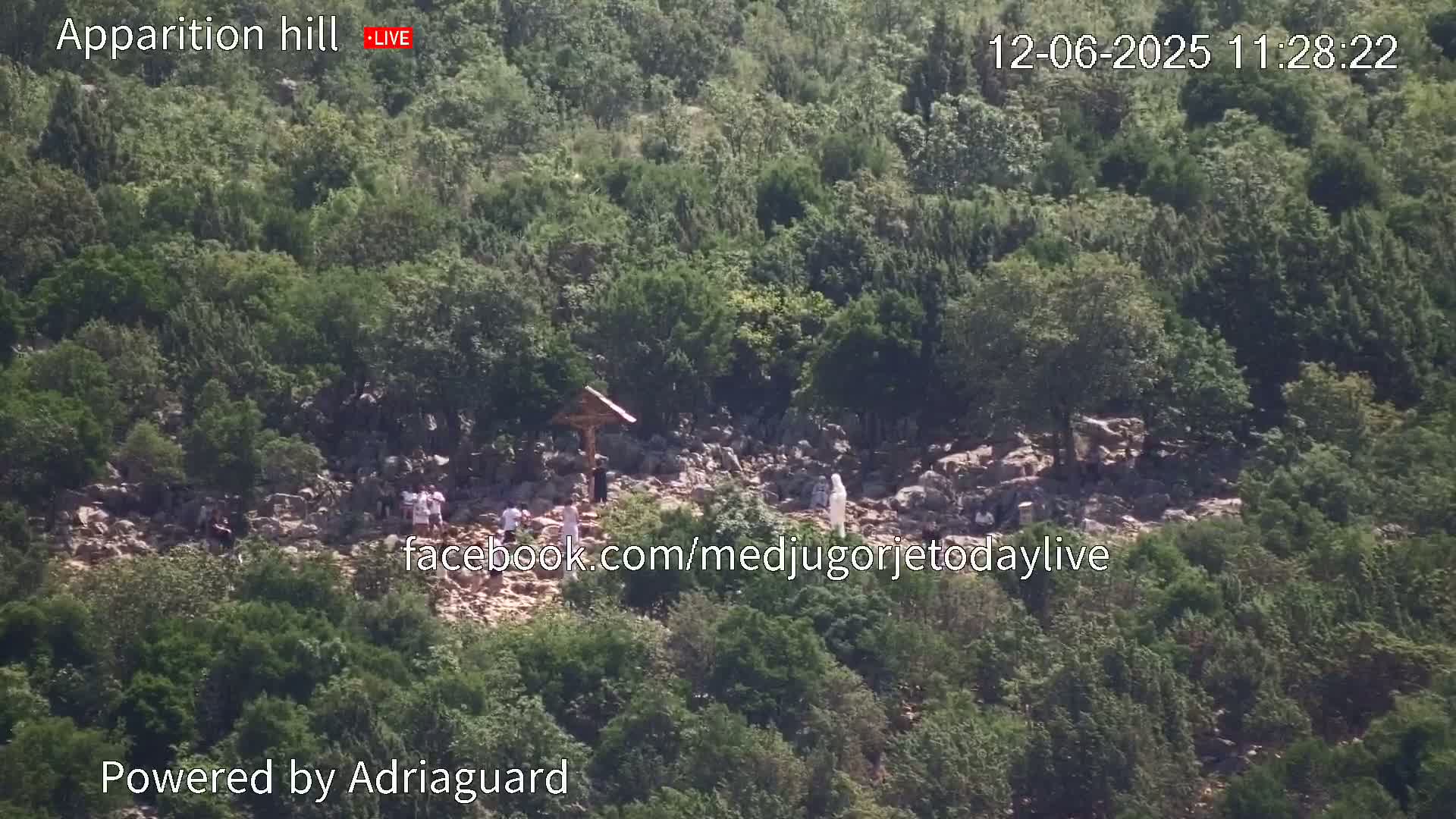 A rocky hillside covered in green vegetation shows a group of people near a wooden cross under a bright, sunny sky.