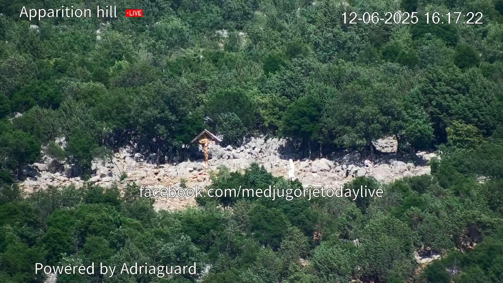A rocky hillside covered in lush green trees displays a crucifix and a statue, under a sunny sky.