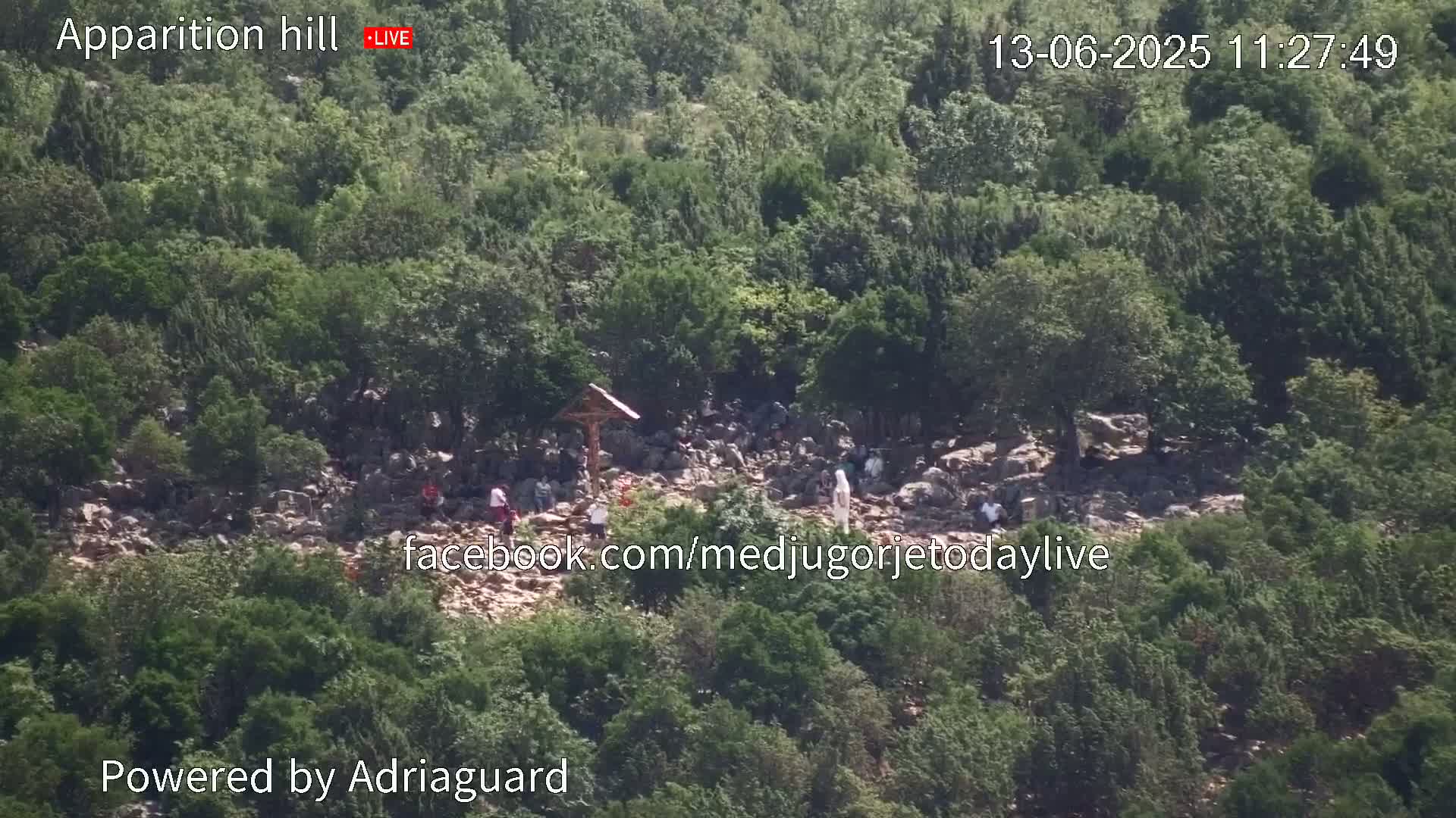 A rocky hillside covered in green vegetation shows a group of people gathered around a cross on a sunny day.