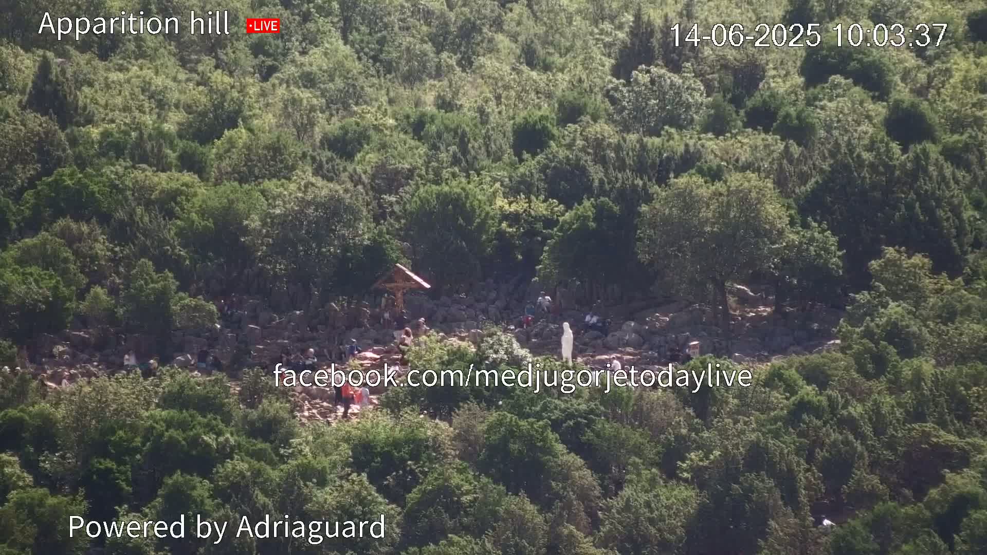 A rocky hillside covered in lush green vegetation shows a gathering of people near a crucifix and a statue.