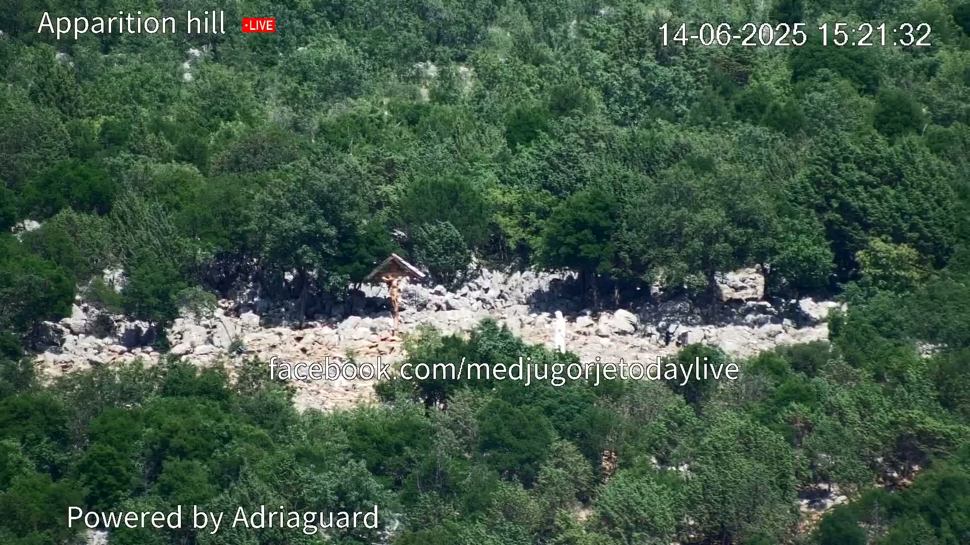 An aerial view shows a rocky hillside covered in lush green trees and shrubs, with a wooden cross visible near its center.