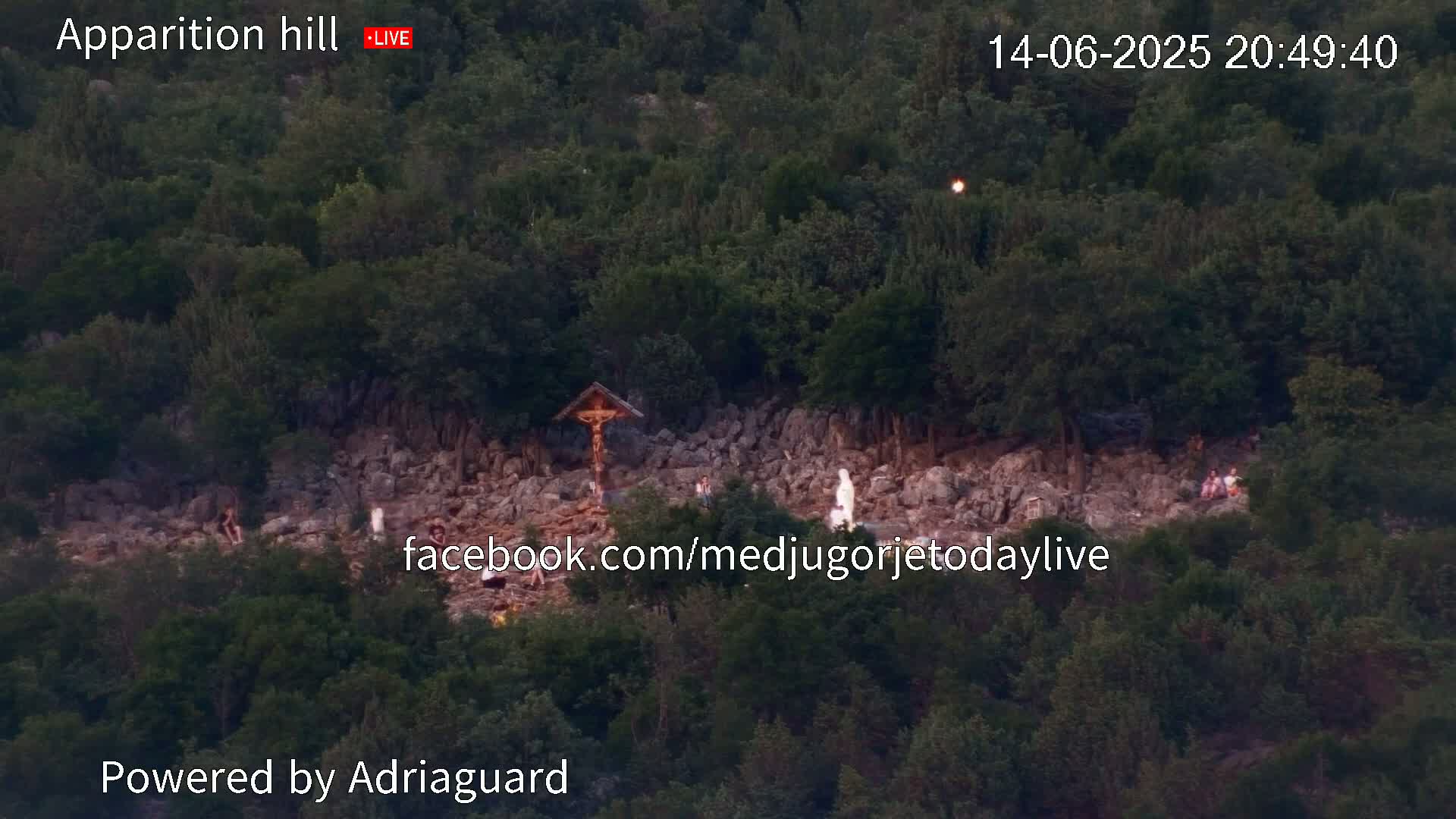 An aerial view shows a rocky hillside covered in lush green trees and shrubs, with a wooden cross visible near its center.