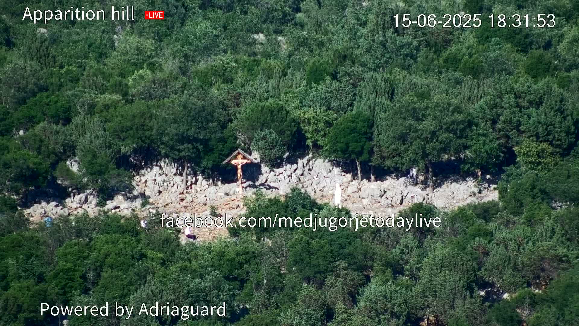 A rocky hillside covered in lush green trees and shrubs features a crucifix and a statue, with a few people visible near the base.