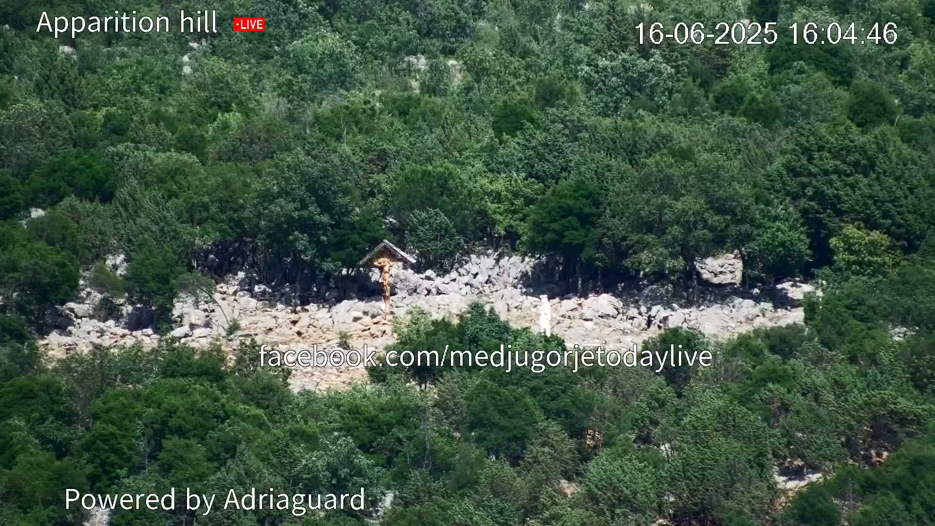 An aerial view shows a rocky area with a crucifix and a small structure nestled amongst lush green trees on a sunny day.