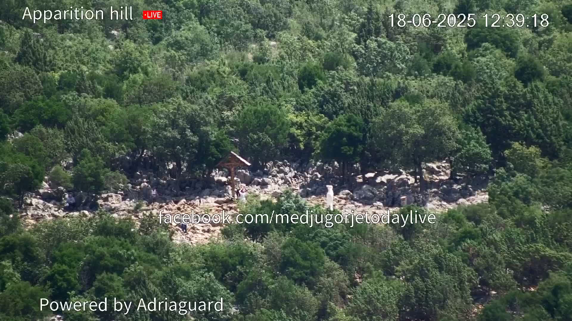 A rocky hillside covered in lush green vegetation shows a few people near a wooden cross.