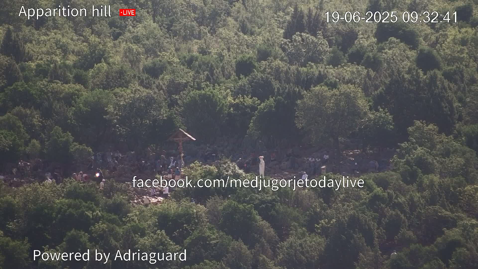 A crowd of people gathers on a tree-covered hillside, near a cross and statue.