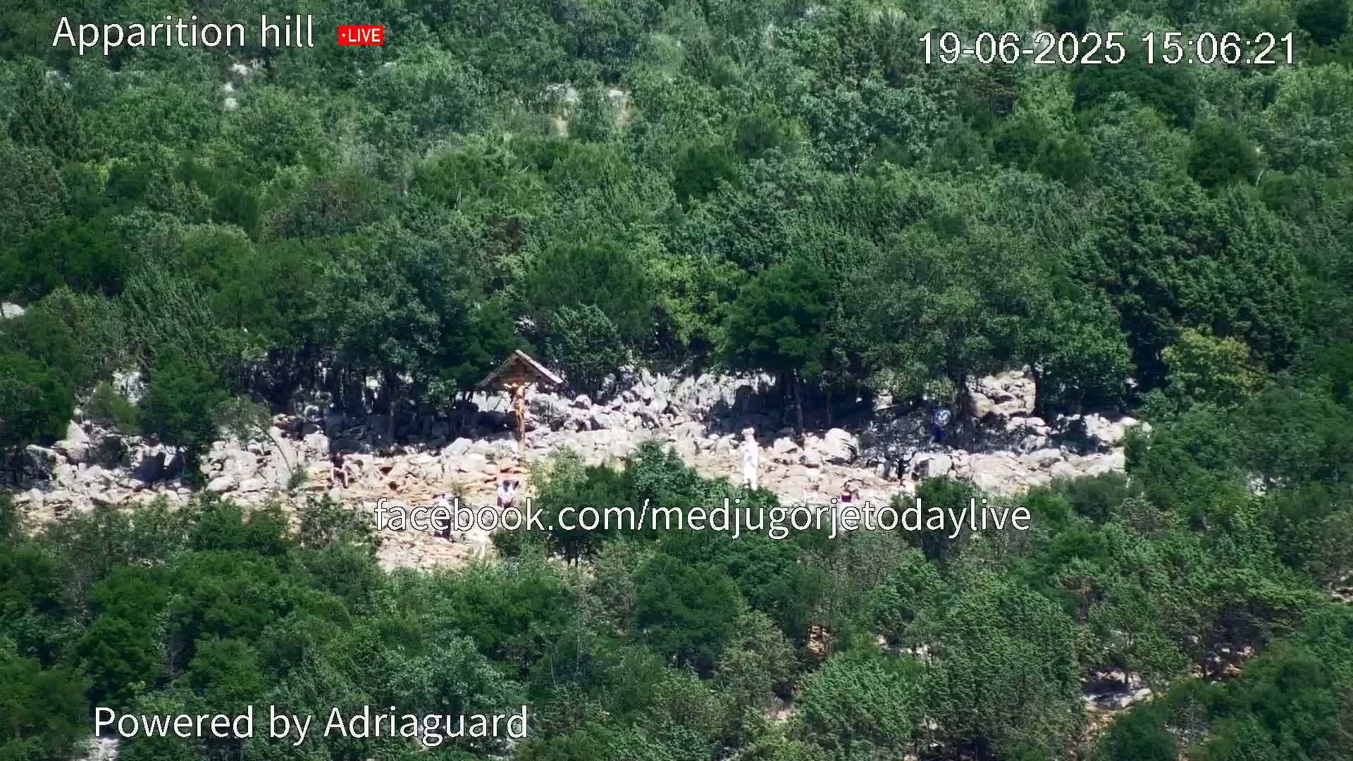 An aerial view shows a rocky hillside covered in lush green trees and shrubs, with several people walking on a path among the rocks near a crucifix.