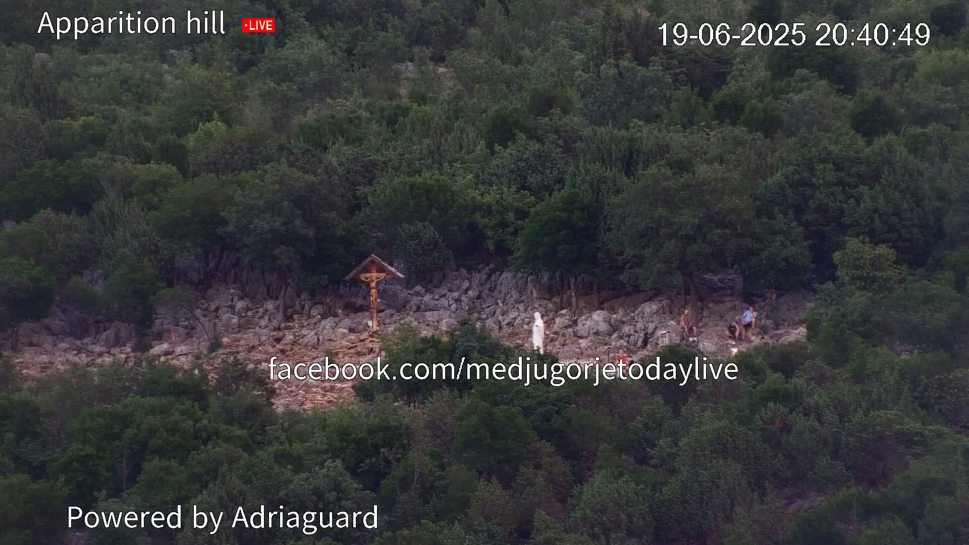 A rocky hillside covered in dense green foliage, with a few people gathered near a crucifix and a statue.