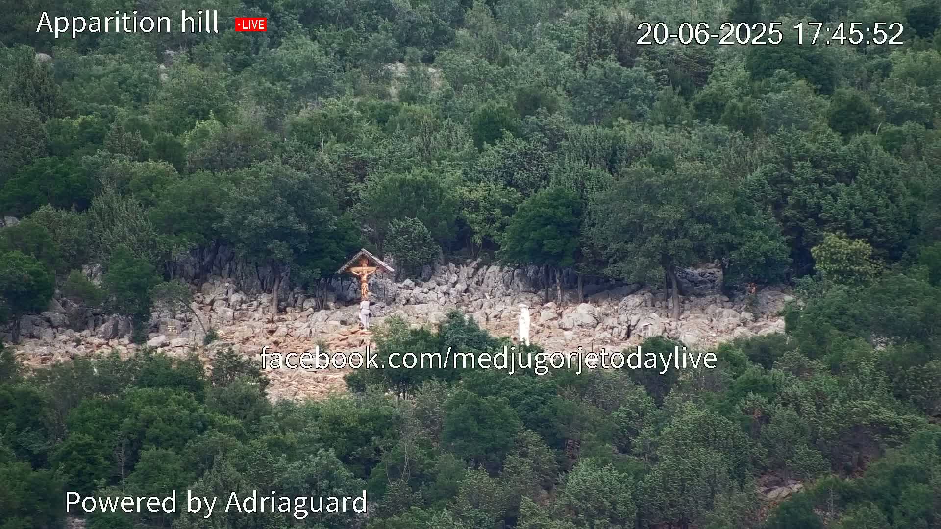 A rocky hillside covered in green trees and shrubs, with a wooden cross and a figure in white visible near the center.