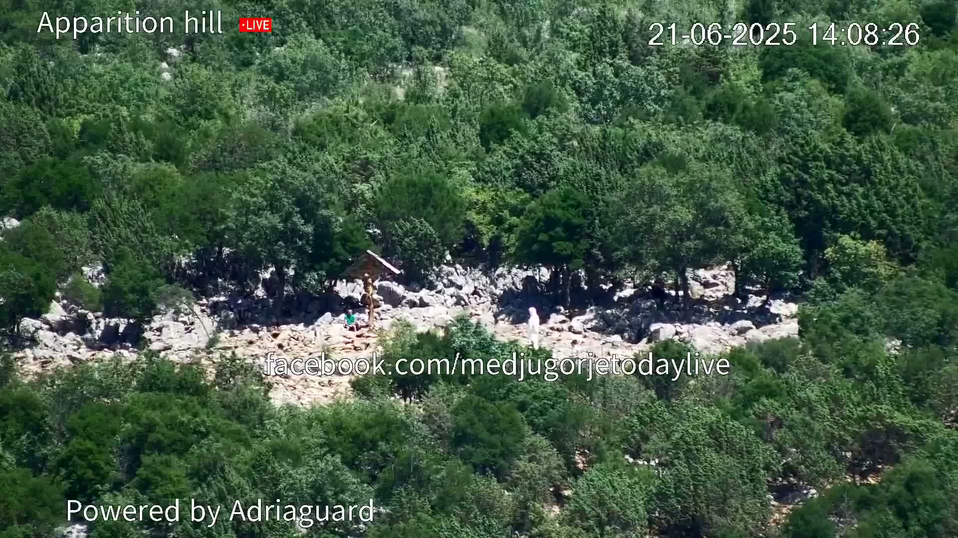 A rocky area with sparse vegetation and a wooden cross is surrounded by lush green trees under a sunny sky.