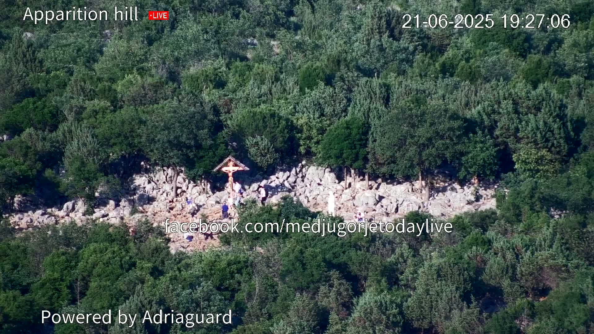 An aerial view shows a rocky hillside covered in green trees and shrubs, with a small group of people gathered around a crucifix.
