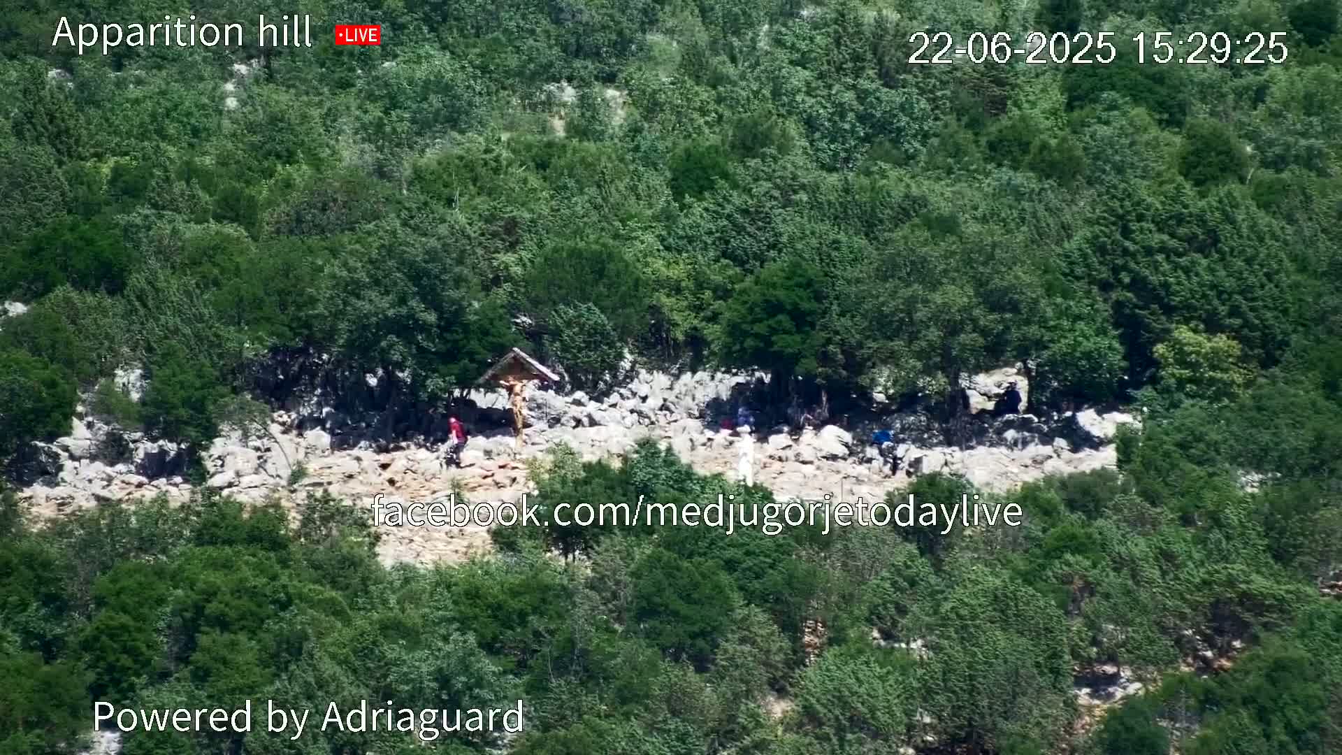 A rocky hillside covered in lush green vegetation shows a group of people gathered around a crucifix.