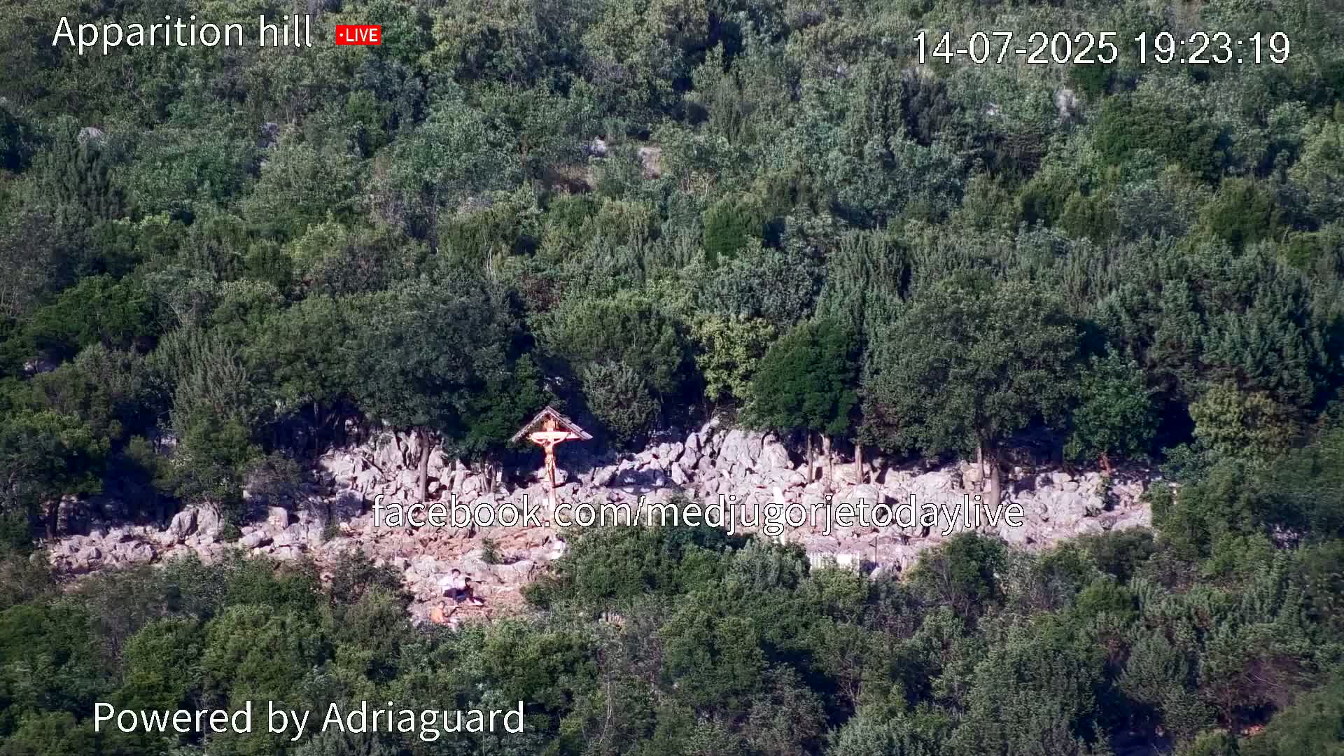 An aerial view shows a rocky hillside covered in lush green trees and shrubs, with a crucifix situated near the center.