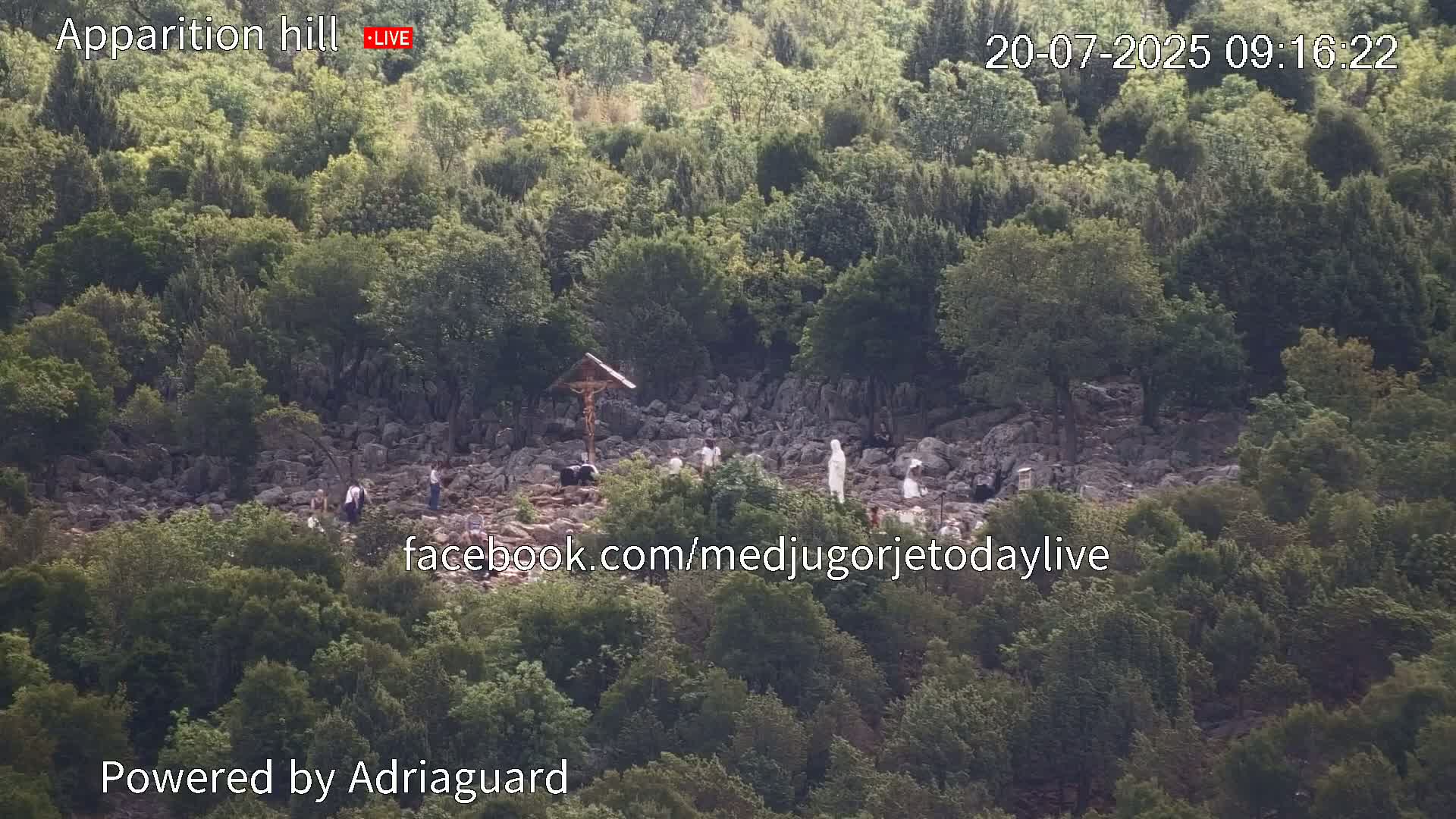 A rocky hill covered in green vegetation, with a few people gathered around a crucifix and a statue.