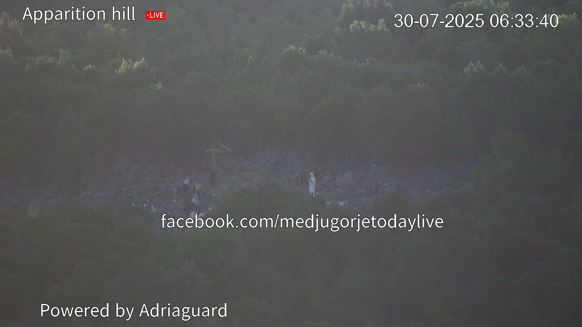 A rocky hill covered in green vegetation, with a few people gathered around a crucifix and a statue.