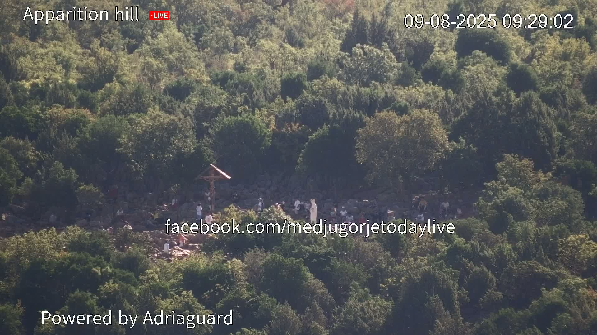 A high-angle view shows a group of people gathered on a rocky hillside covered in green trees under a bright, sunny sky.