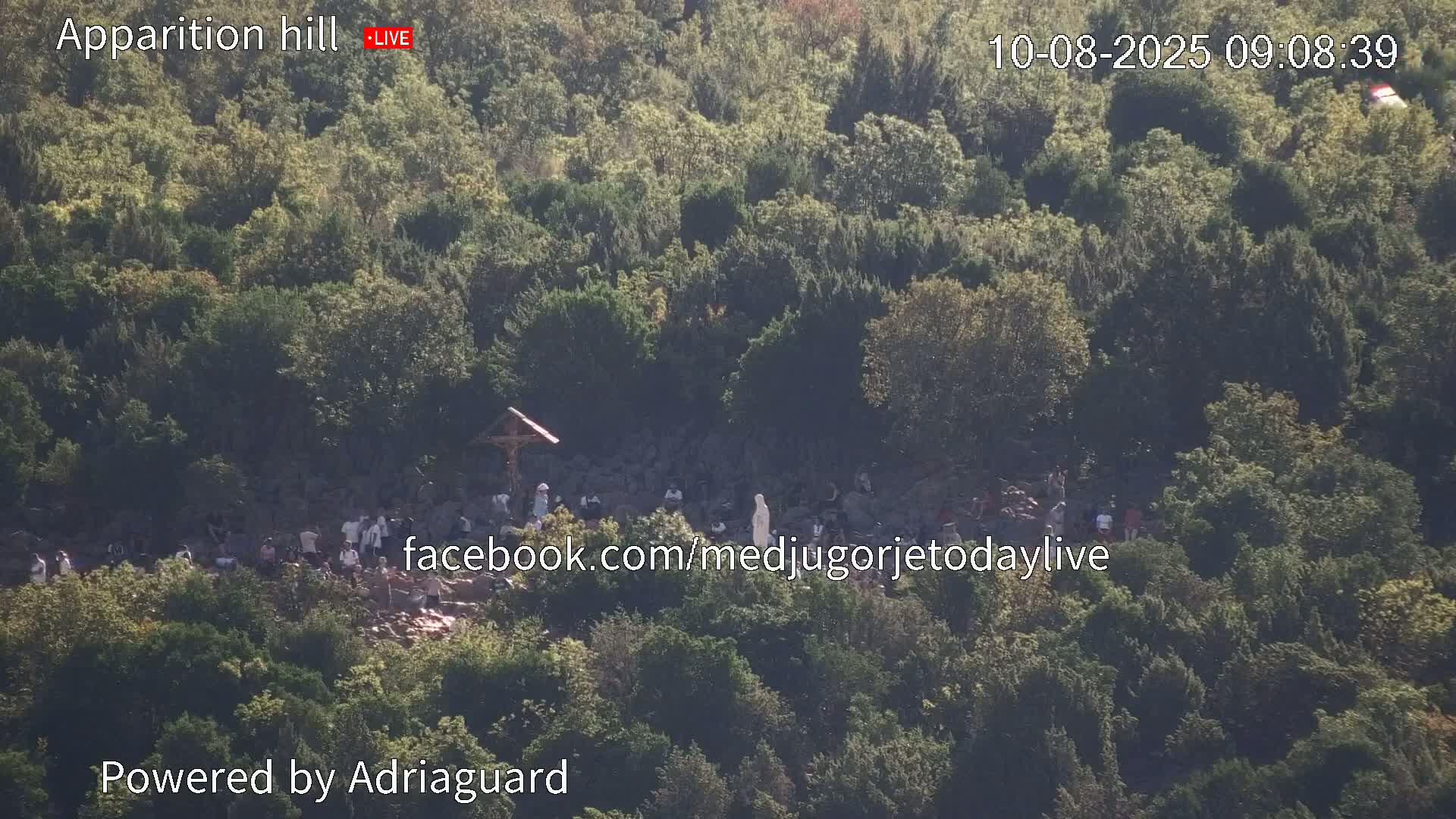 A crowd of people gathers on a rocky hillside covered in green trees under a bright, sunny sky.