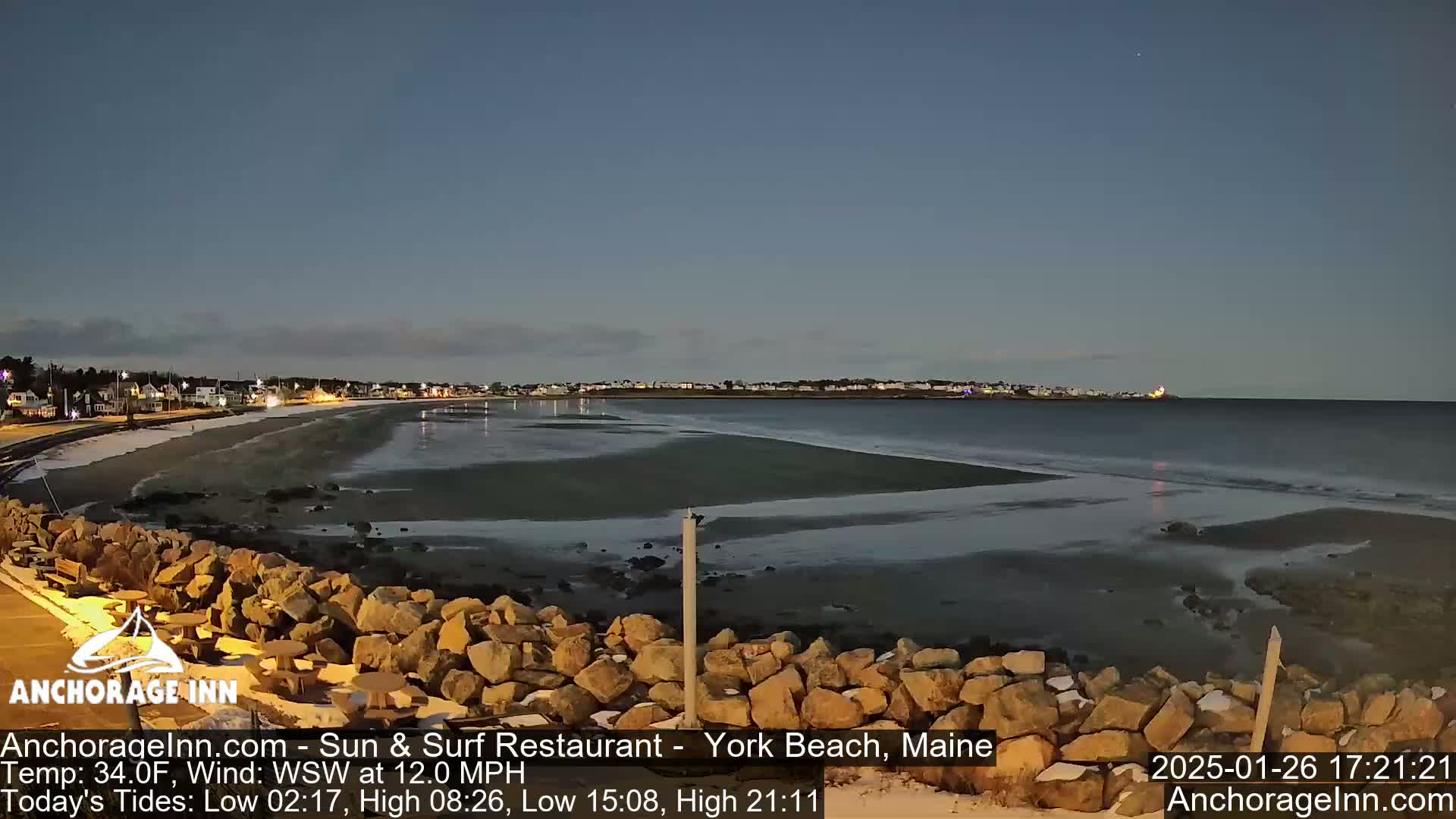 A nighttime view of a partly icy beach with a calm ocean and a town illuminated in the distance under a twilight sky.