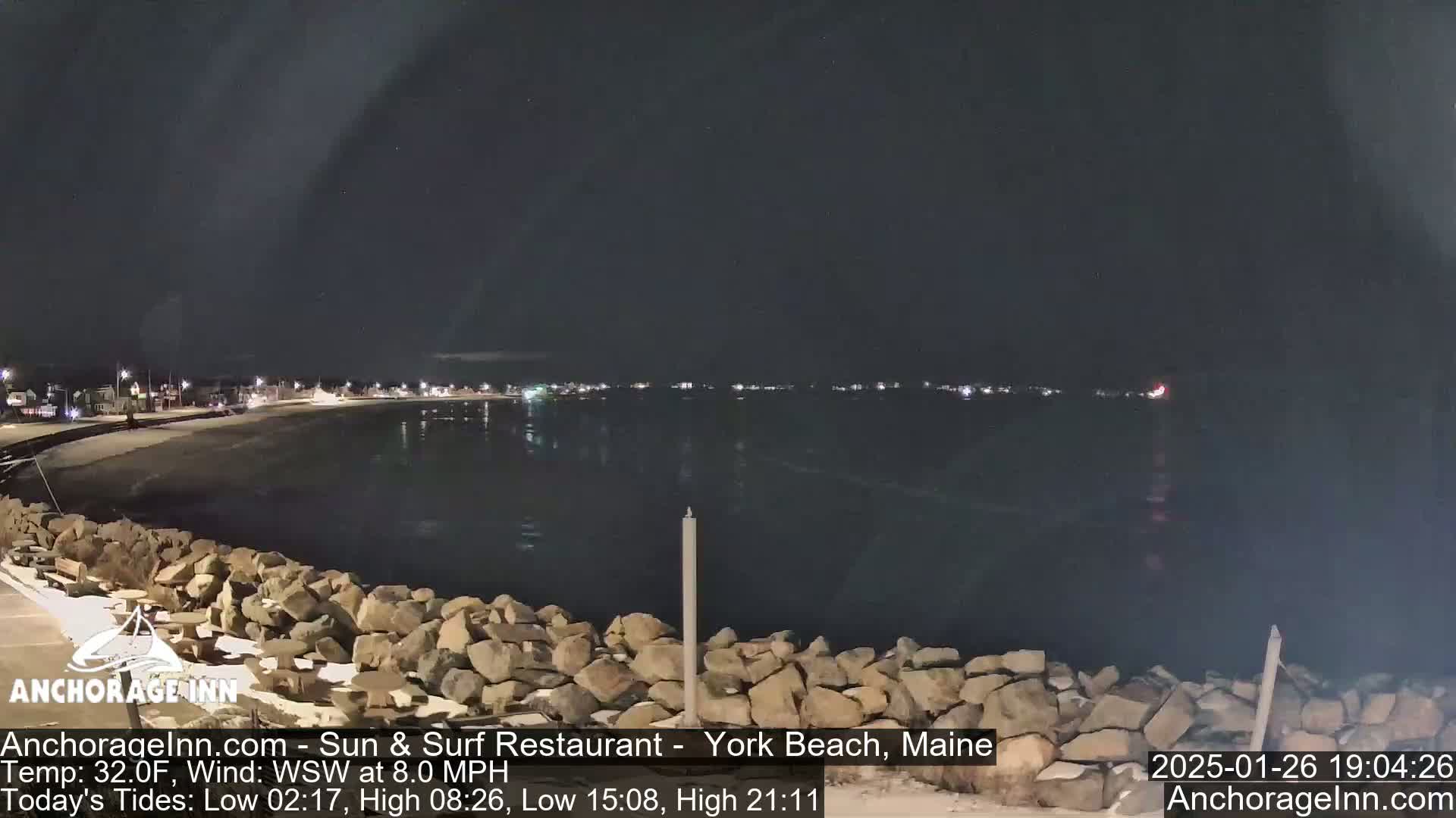 A nighttime view of a calm body of water with a shoreline of dark rocks under a clear, dark sky.