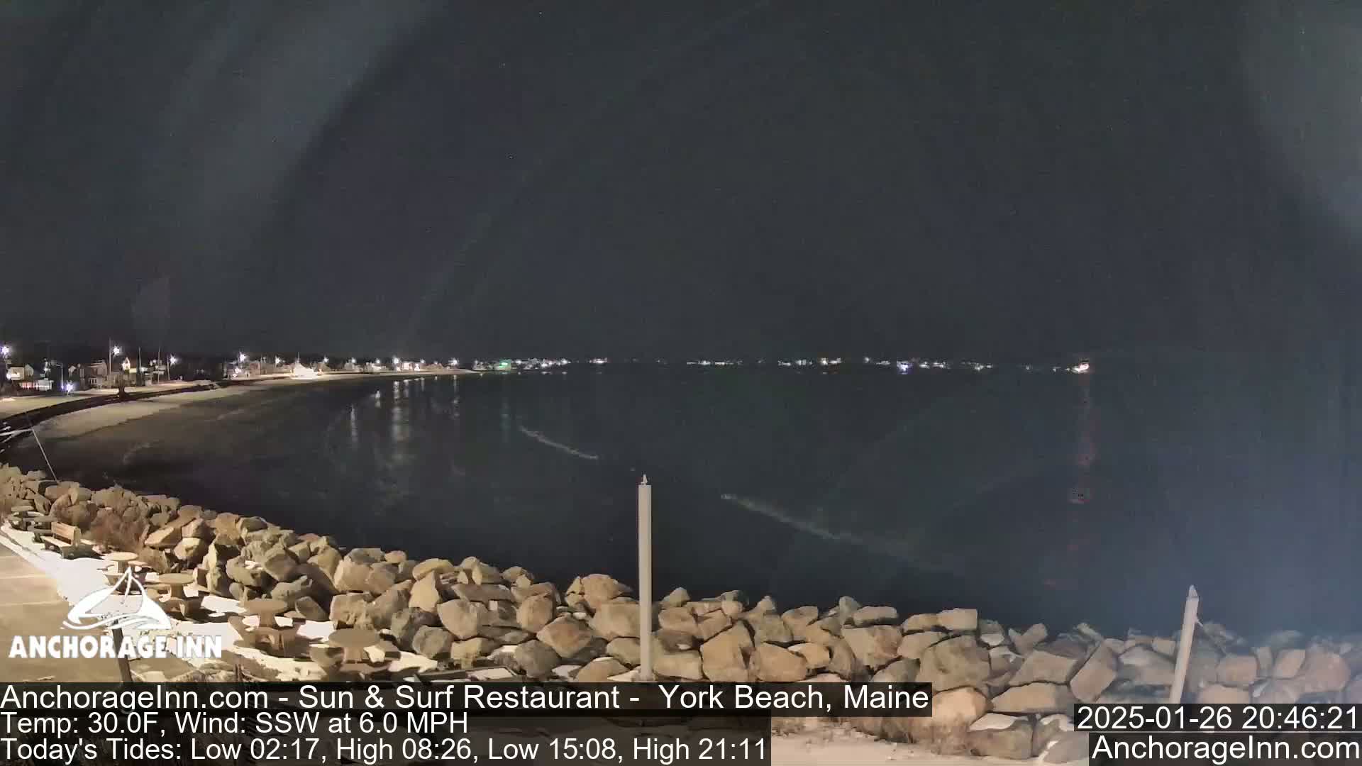 A nighttime view of a calm, dark body of water next to a rocky shoreline, with a town faintly lit in the distance under a clear sky.