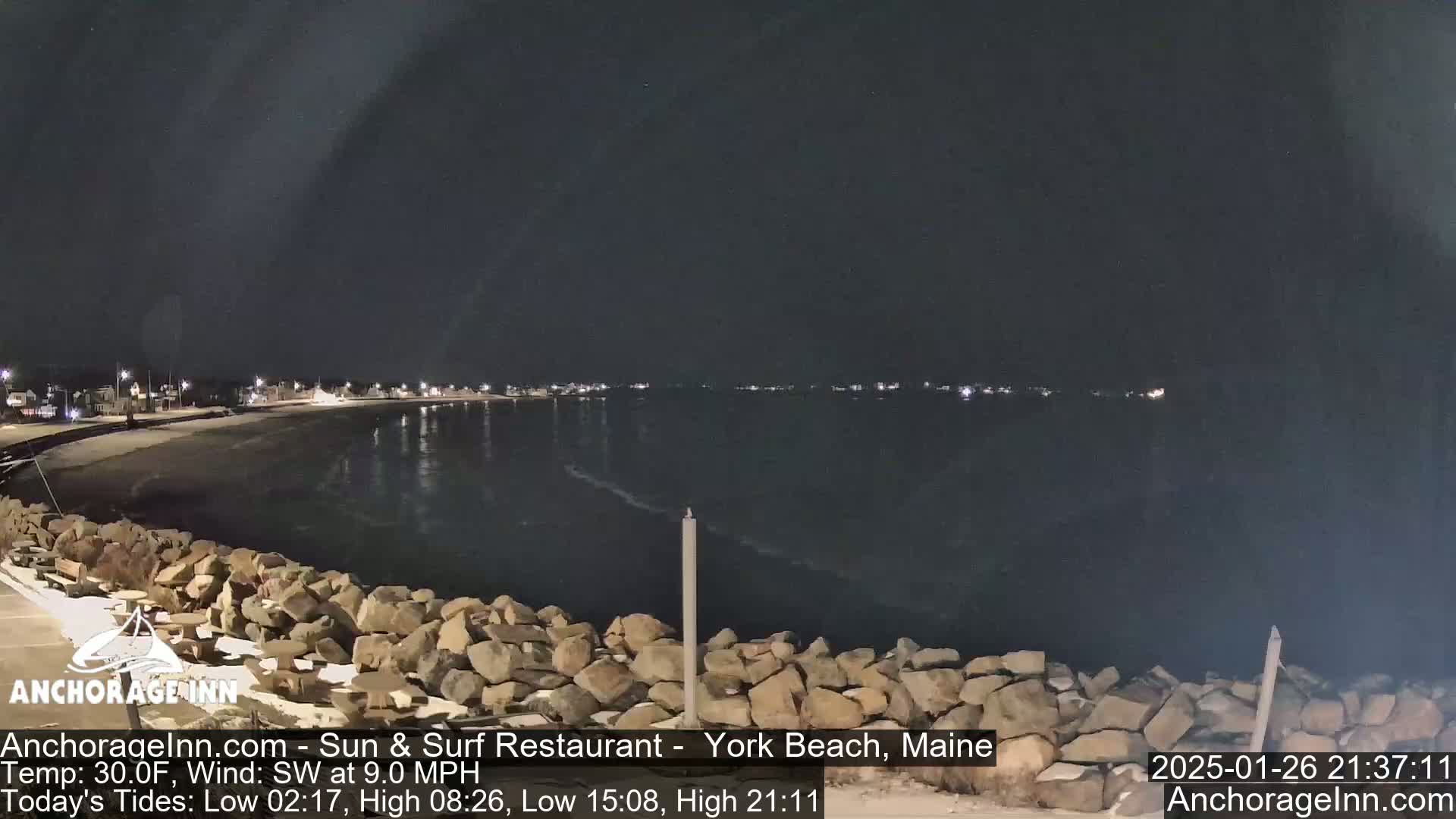 A nighttime view of a calm, dark body of water next to a rocky shoreline with faintly lit buildings in the distance under a clear sky.