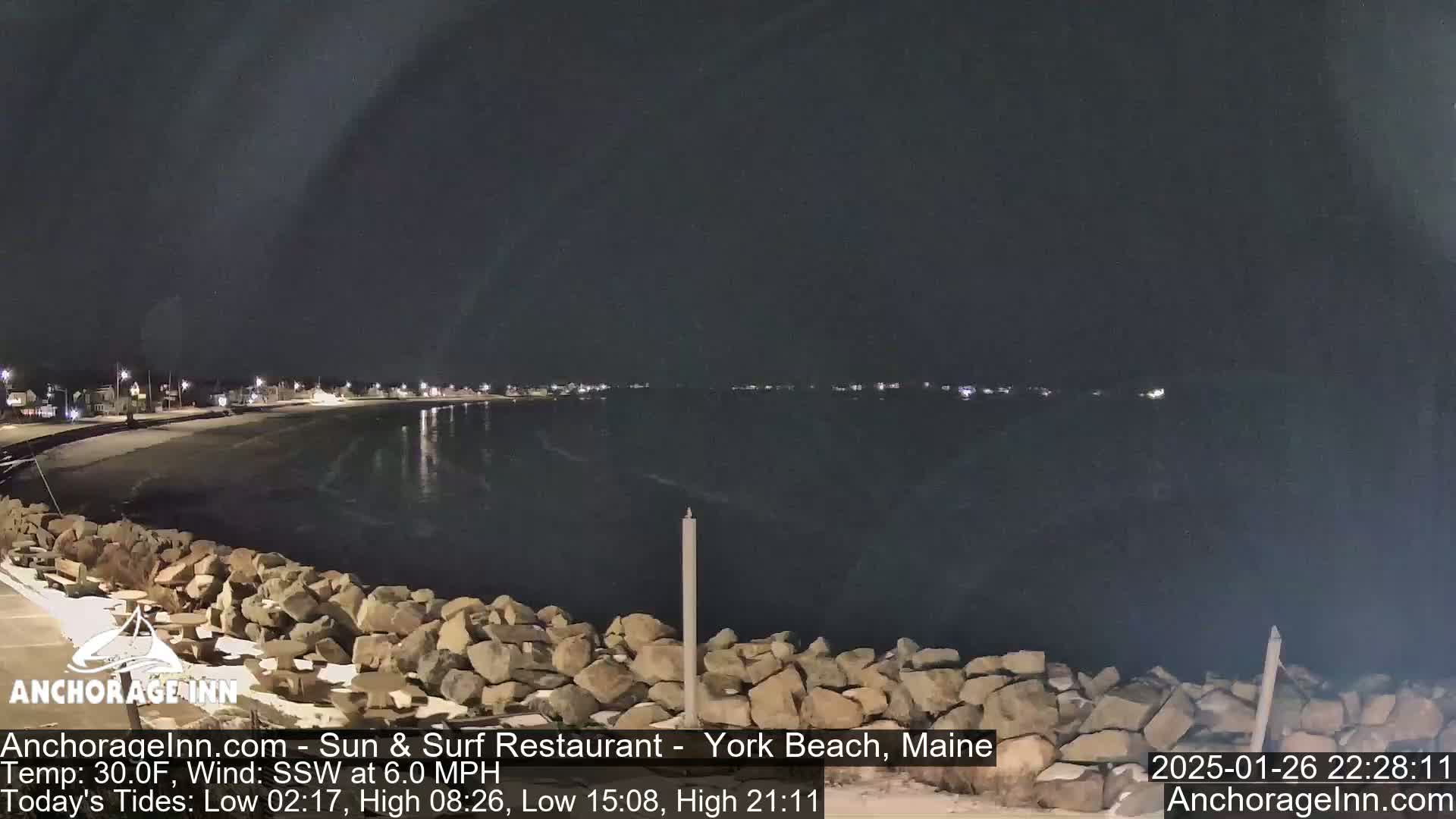 A nighttime view of a calm, dark ocean lapping against a rocky shore, with a town faintly lit in the distance under a clear, dark sky.