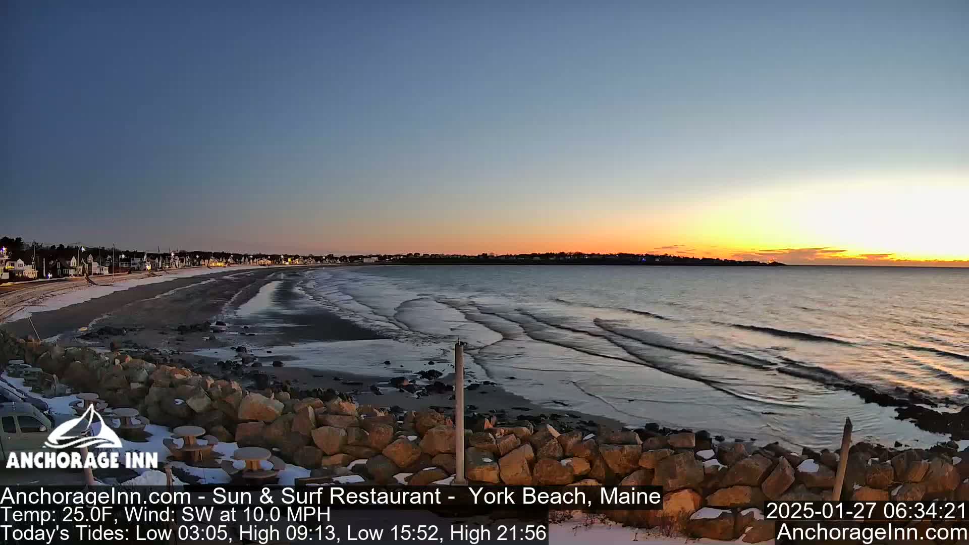 A sunrise over a calm ocean beach with gentle waves rolling onto a snow-dusted shore, backed by a line of houses.