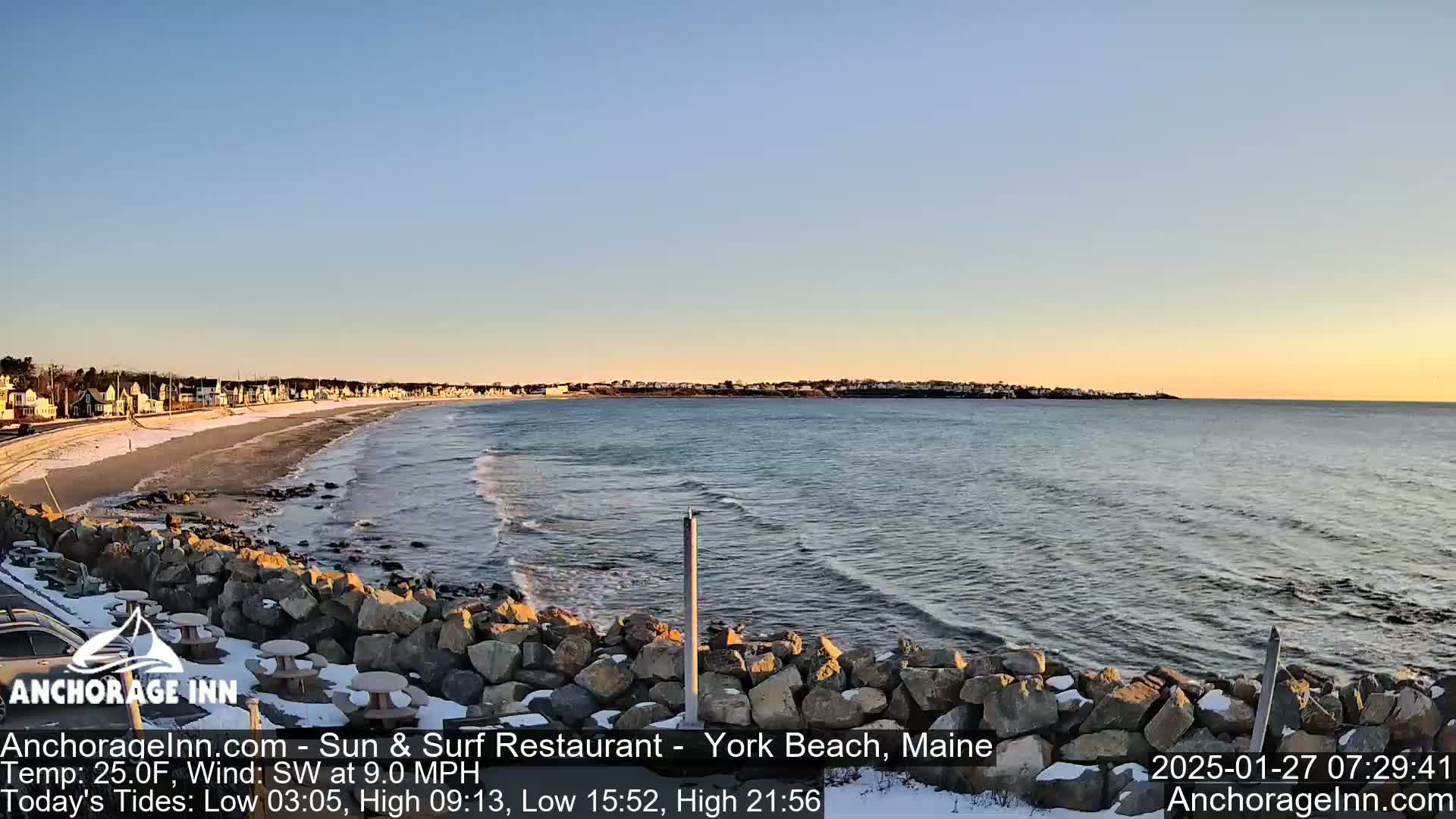 A sandy beach with light wave action and a small amount of snow is visible near a town, all under a clear, light-orange sunset sky.
