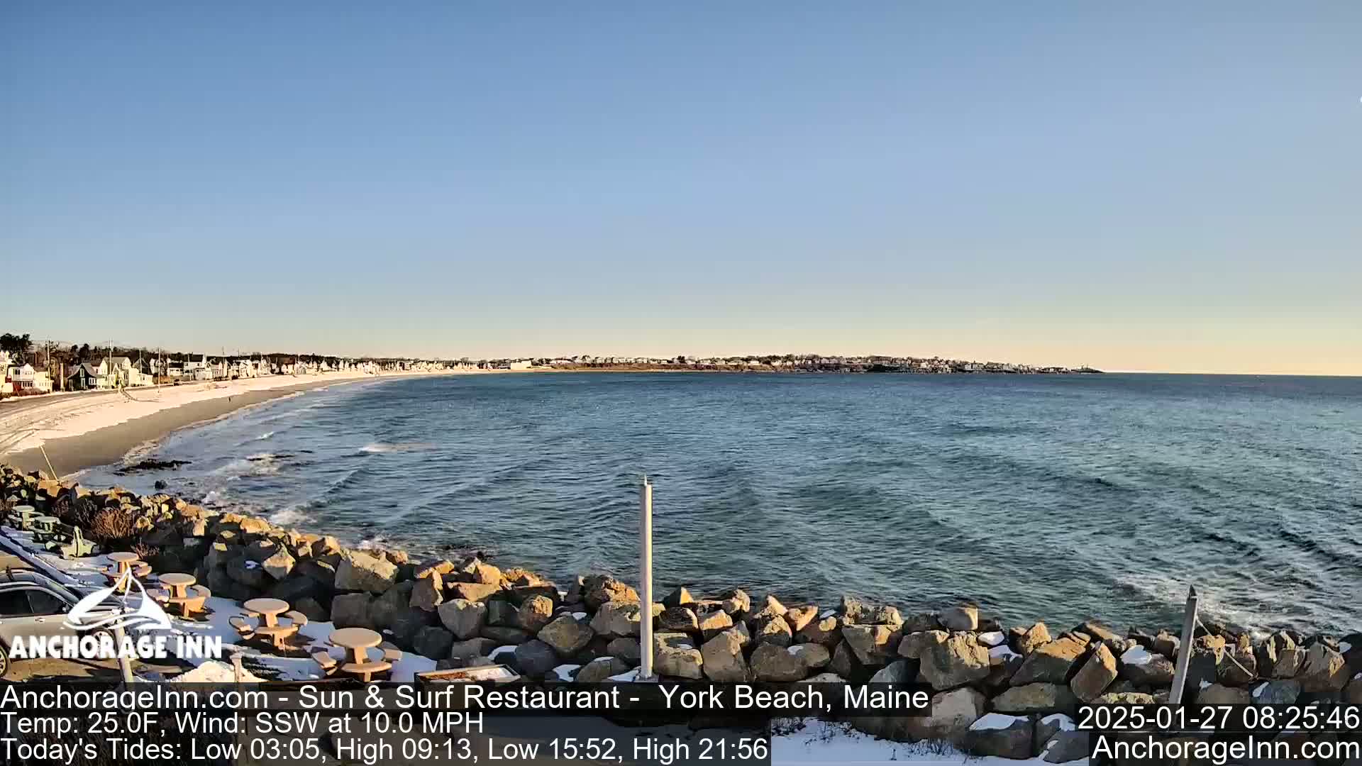A partially snow-covered rocky shoreline overlooks a calm ocean with a residential area visible in the distance under a clear blue sky.