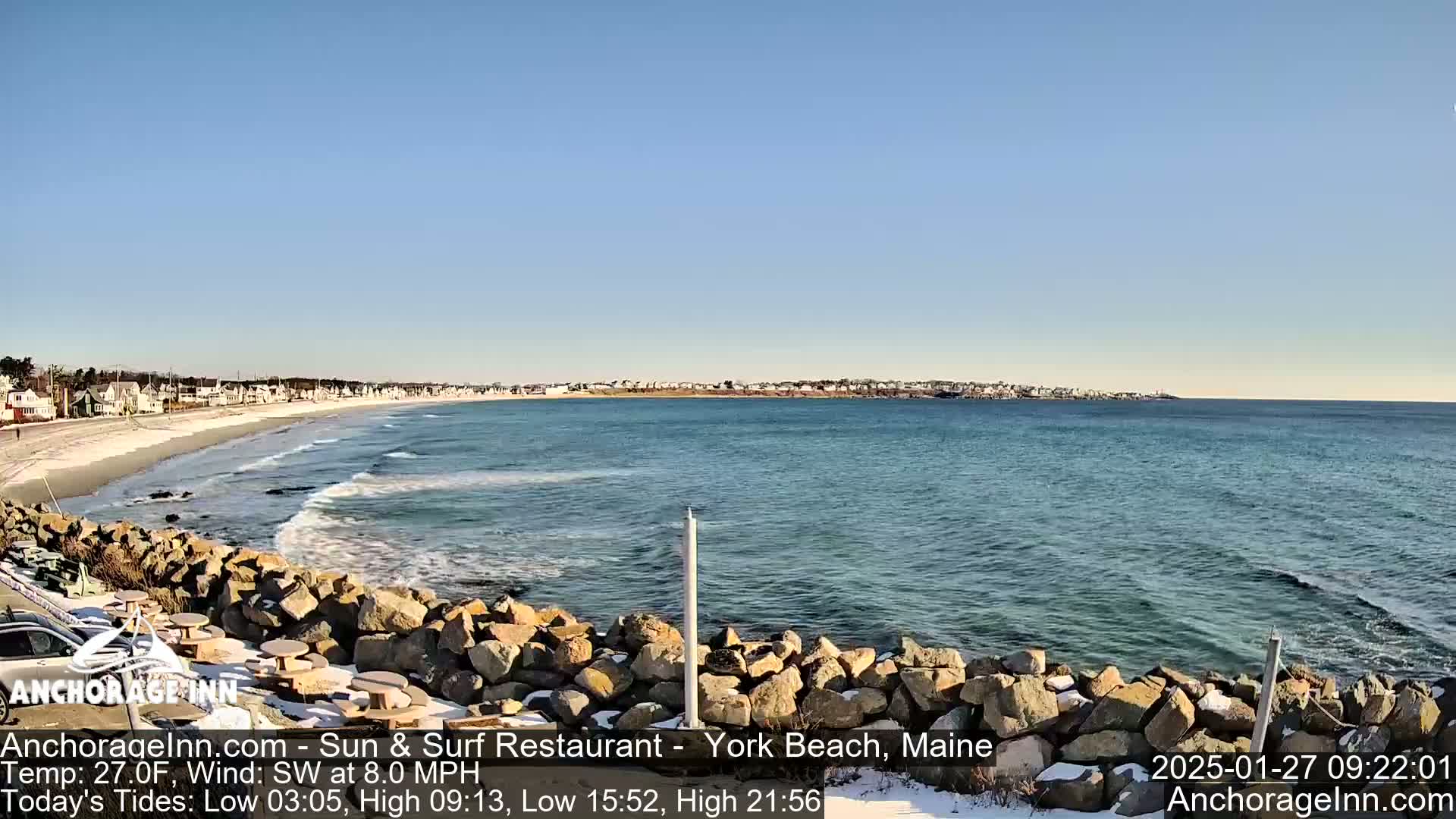 A sunny day at a rocky beach shows a calm ocean with small waves, a sandy shore with snow, and houses in the distance.