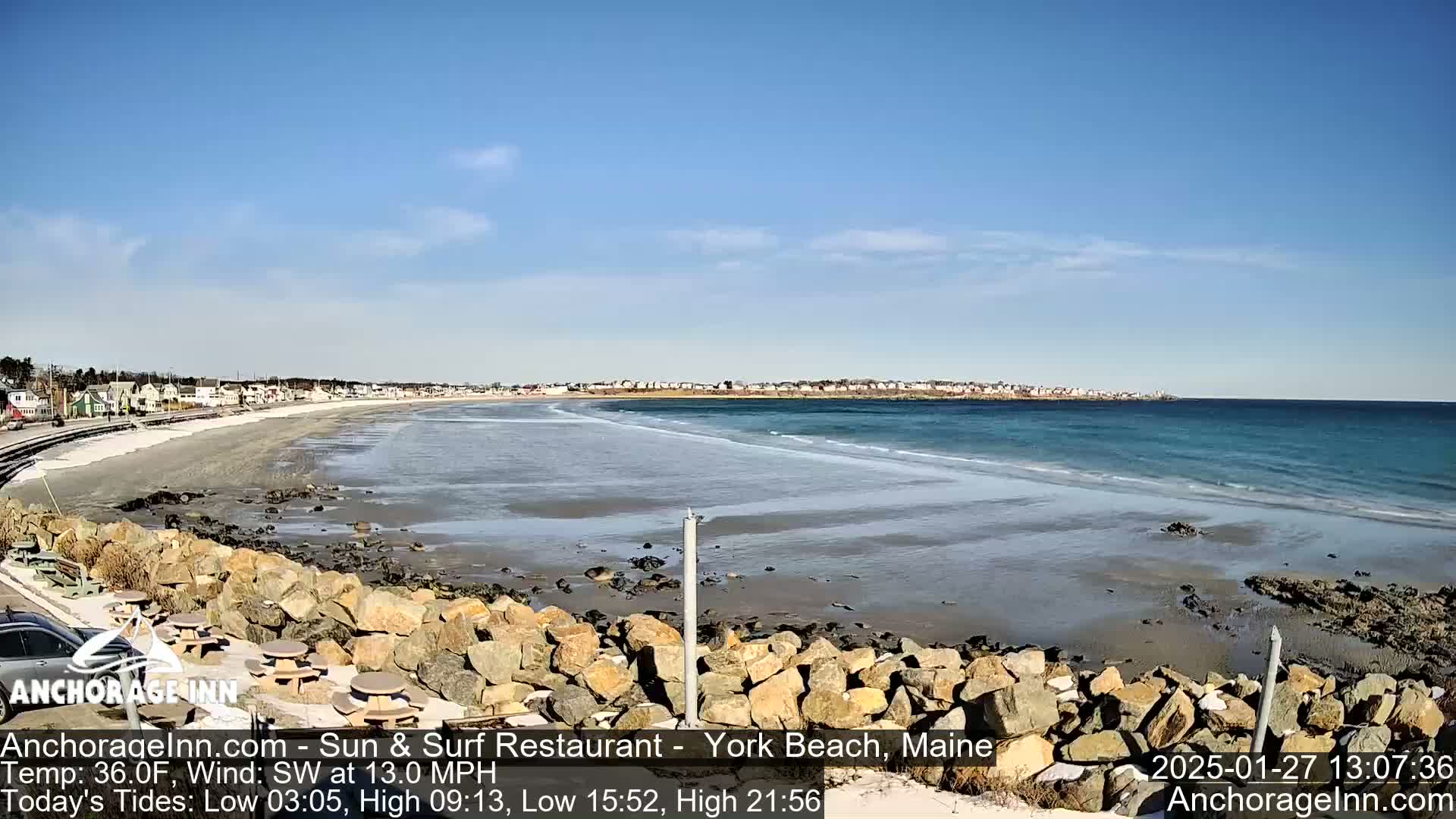 A sandy beach with a calm ocean and a clear blue sky stretches out from a rocky shoreline; houses and buildings are visible in the distance.
