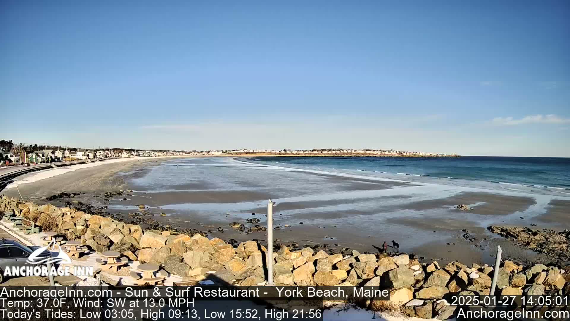 A mostly sunny day shows a sandy beach with some ice patches, small waves, and a rocky shoreline, with a town visible in the distance.
