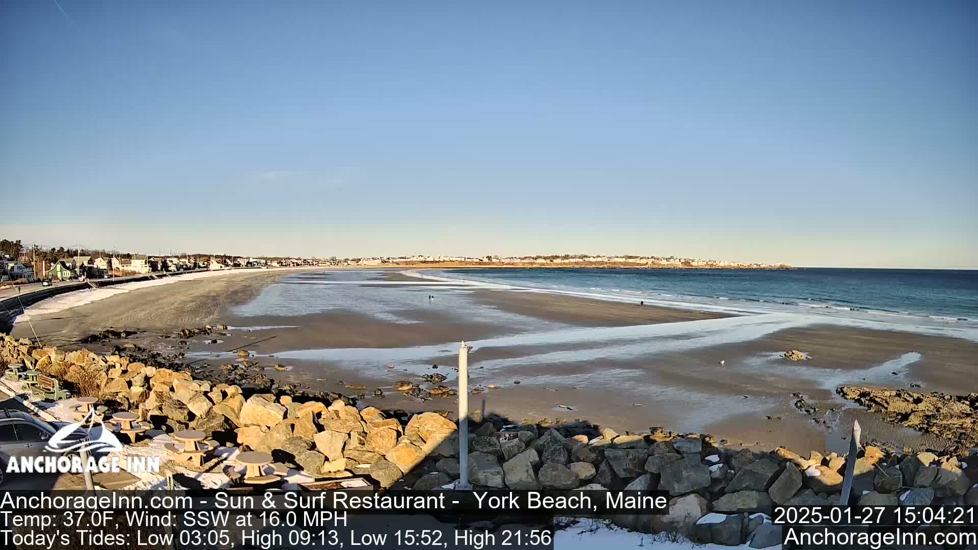 A partially icy sandy beach stretches along a coastline with a town visible in the distance under a clear blue sky.