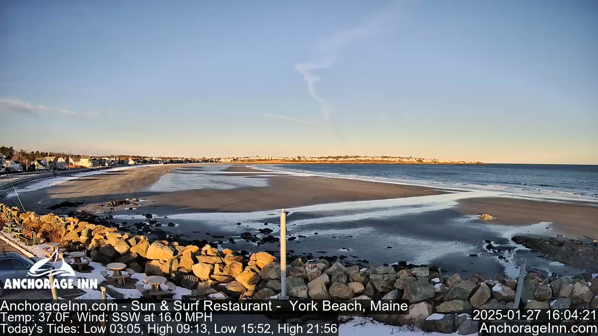A sandy beach with patches of ice and snow stretches out to a calm ocean under a clear, mostly sunny sky.