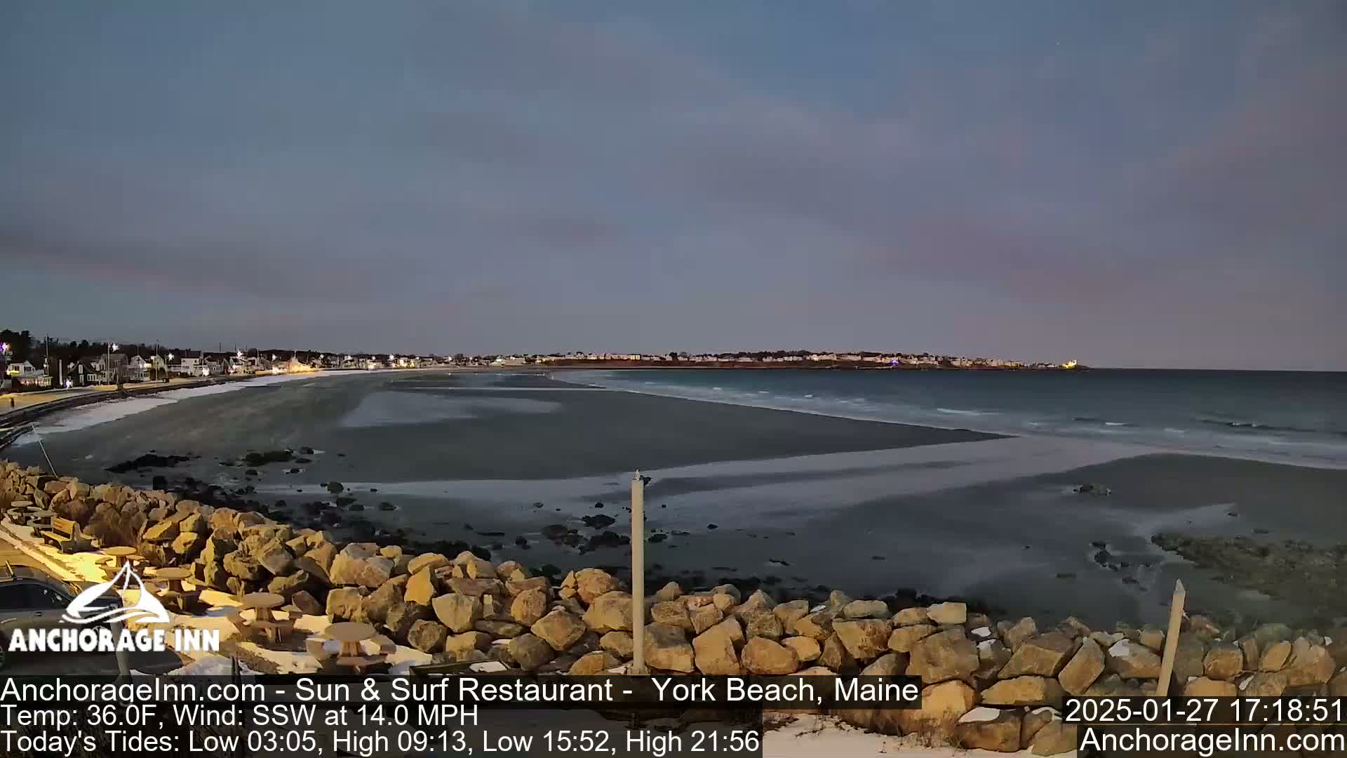 A dark, mostly clear evening at the beach shows a calm ocean with small waves lapping a sandy shore, backed by a town with lights illuminated against the twilight sky.