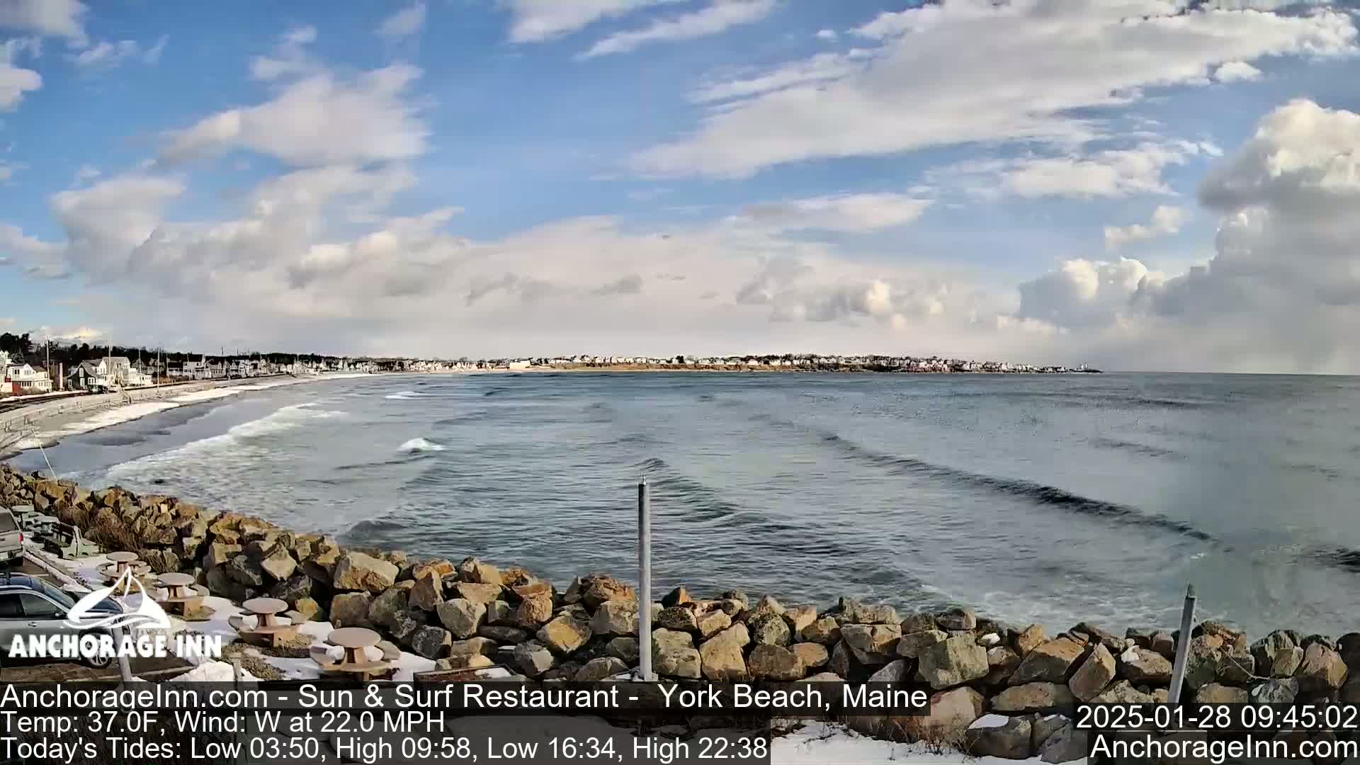 A partly cloudy day shows a rocky shoreline with a beach and ocean waves, some light snow is visible on the ground.