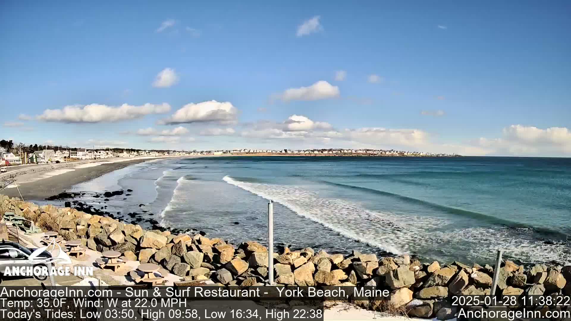 A rocky outcrop overlooks a beach with gentle waves rolling in under a partly cloudy blue sky.