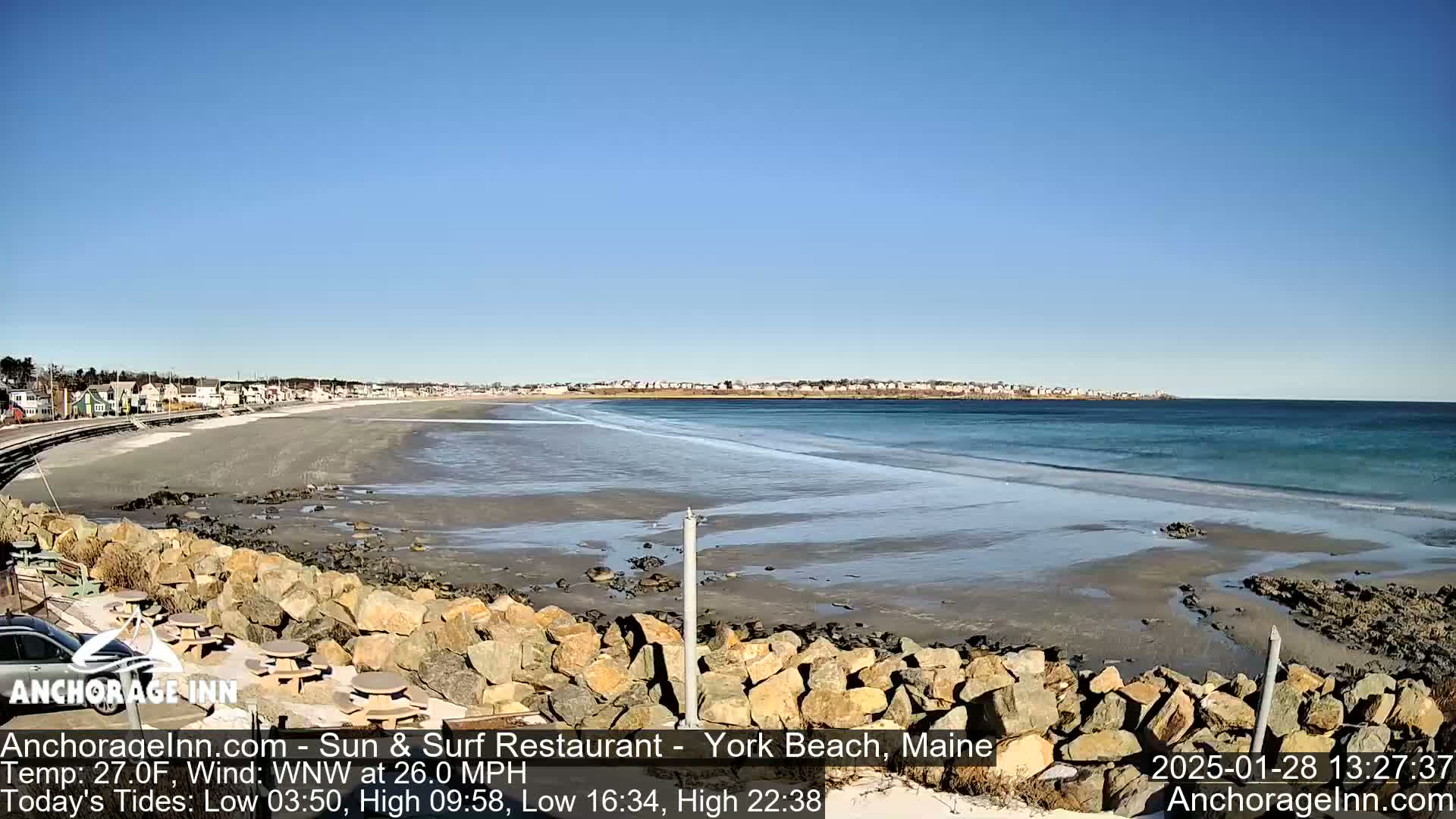 A sandy beach with a rocky shoreline and calm ocean waves stretches out to a distant town under a clear blue sky.
