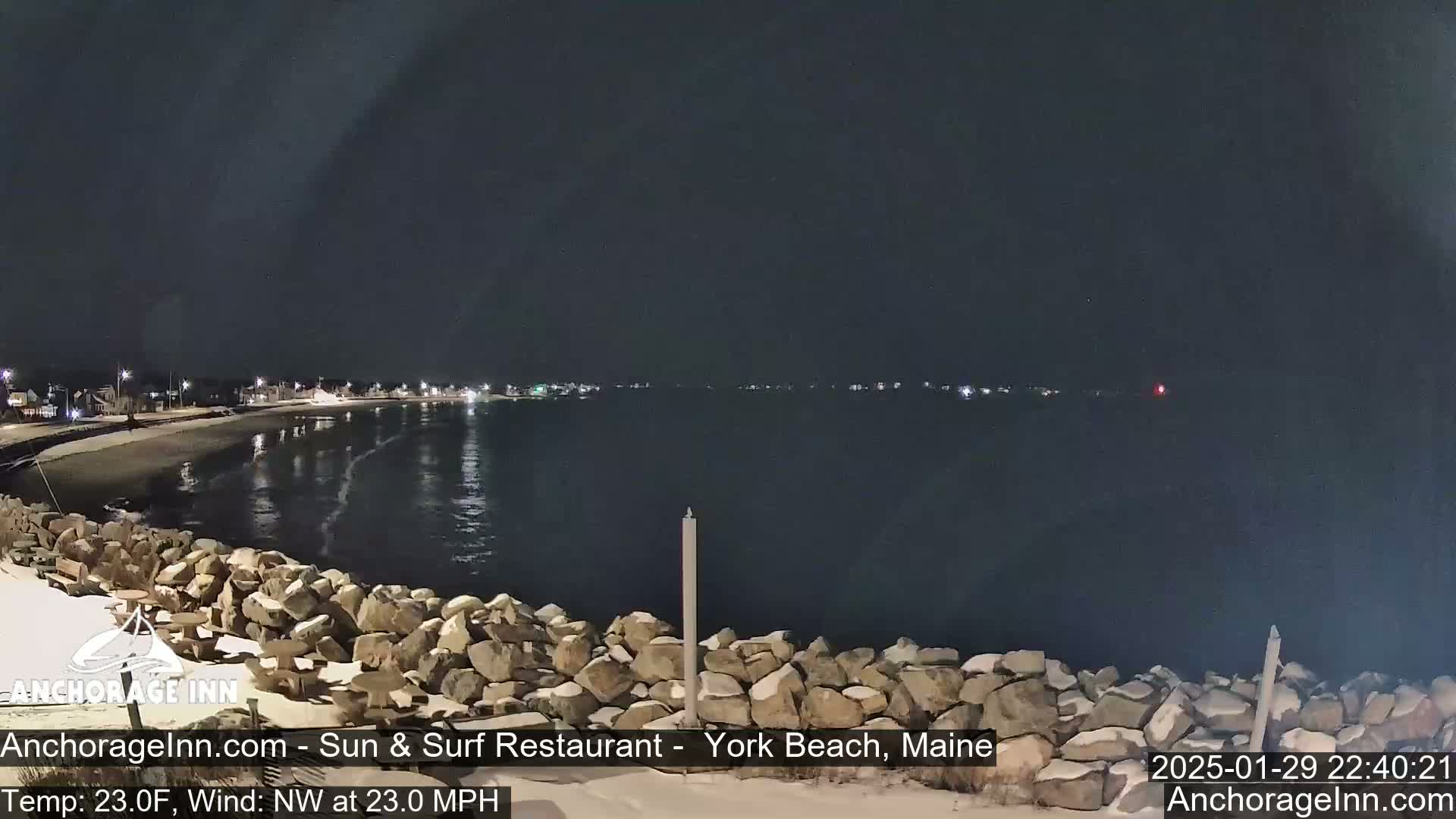 A nighttime view of a rocky shoreline and calm dark water, with a town faintly lit in the distance under a clear, dark sky.