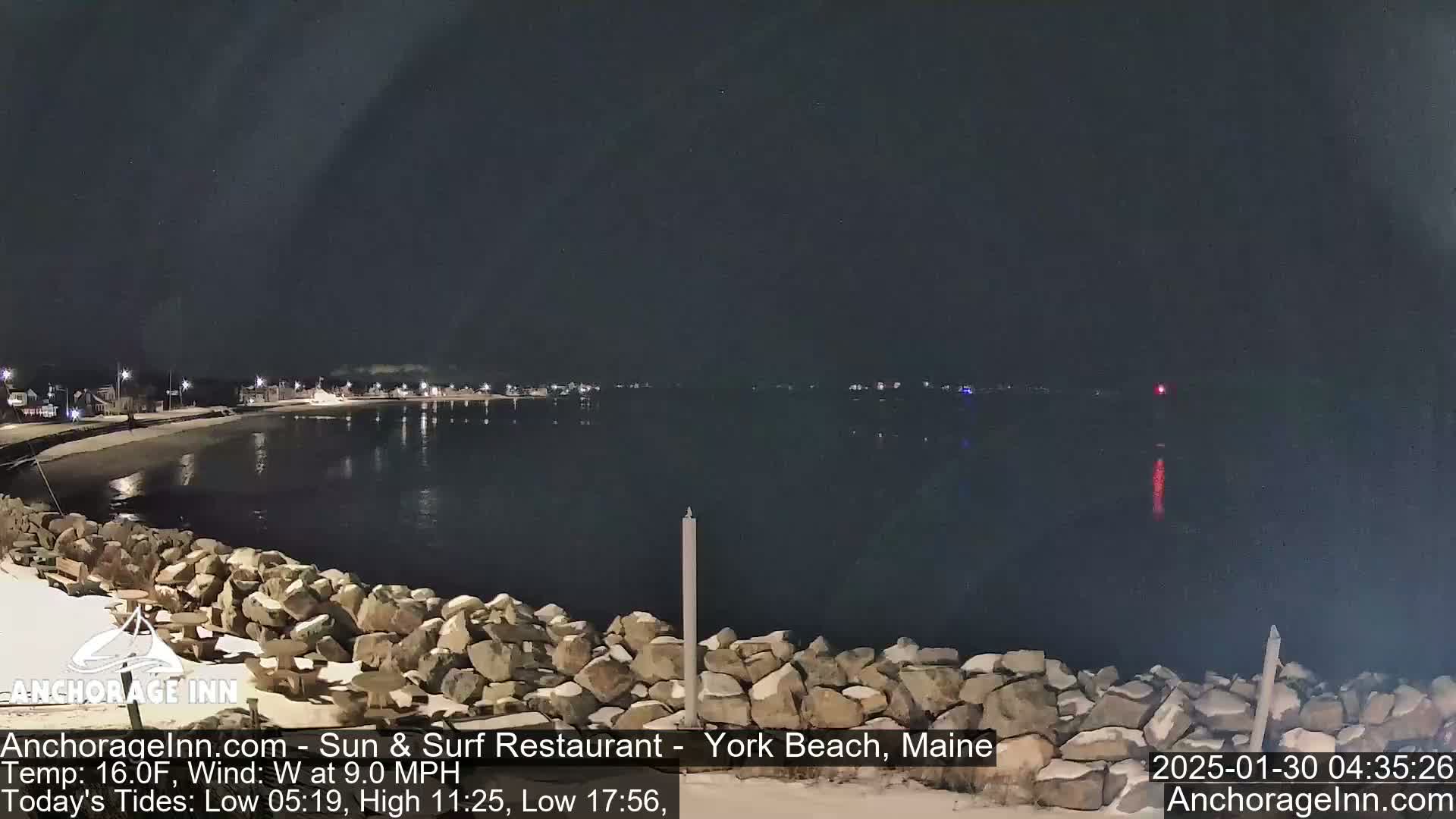 A nighttime view of a rocky shoreline and calm water, with distant lights visible across the water under a dark sky.