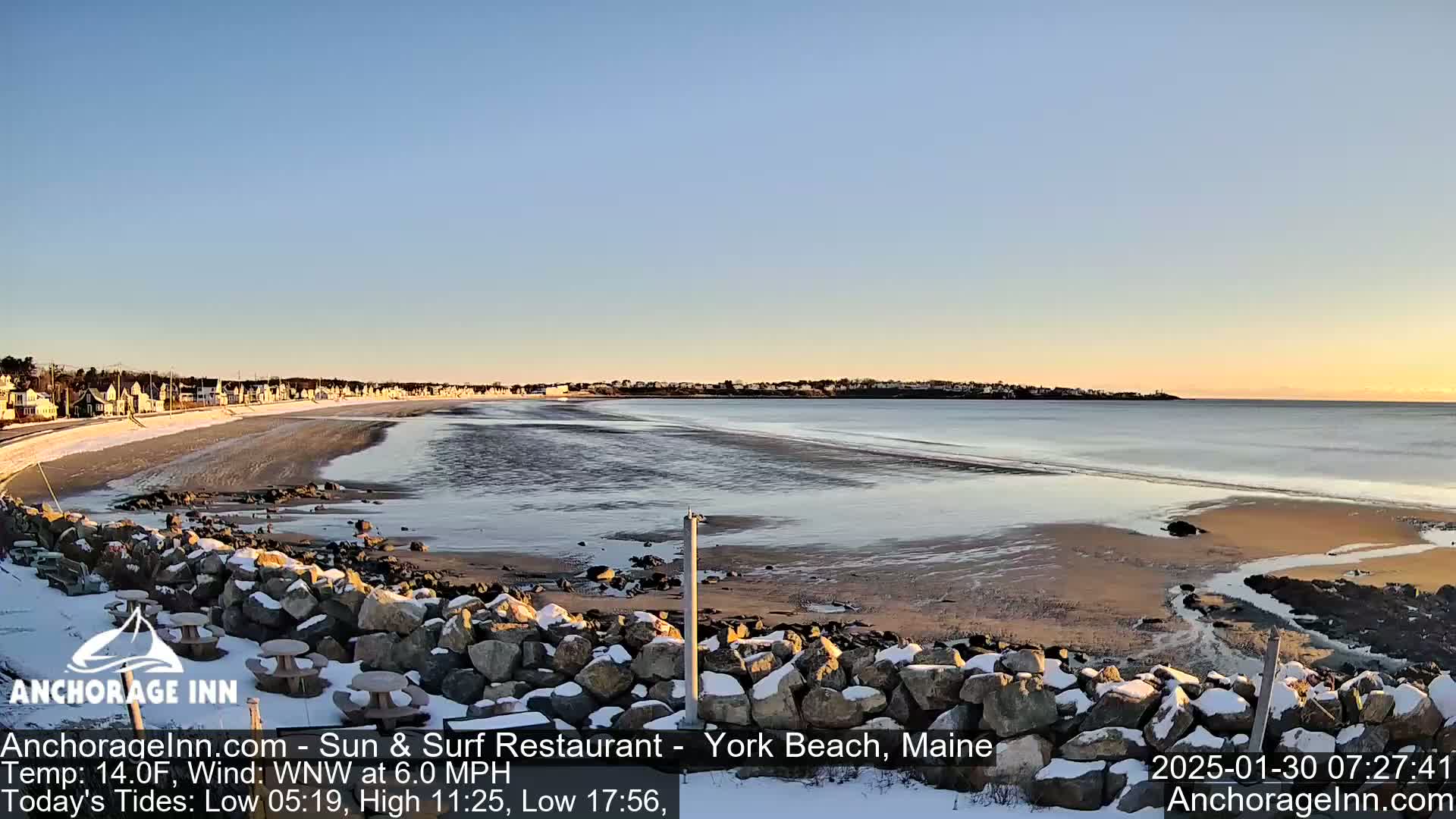 A snowy beach with calm ocean waters and a row of houses in the distance under a clear, sunset sky.