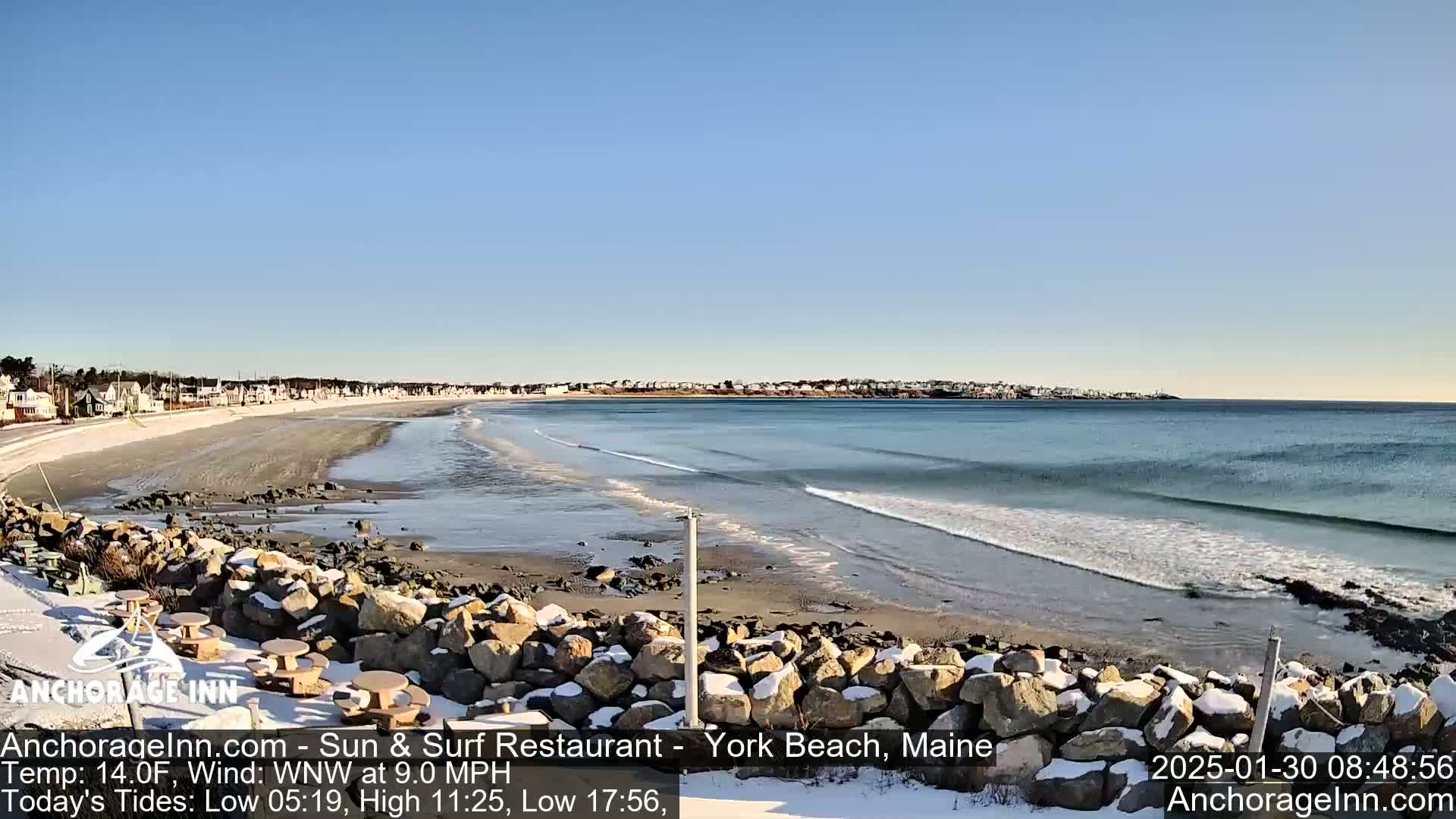 A snow-dusted beach with rocky outcroppings and picnic tables overlooks a calm ocean with gentle waves under a clear blue sky.