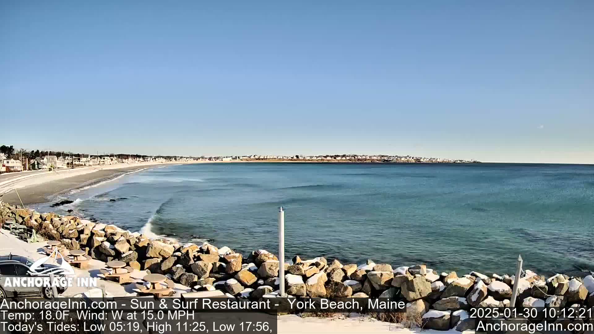 A sandy beach with a rocky shore and calm ocean waves under a clear blue sky; some snow is visible on the rocks and beach.