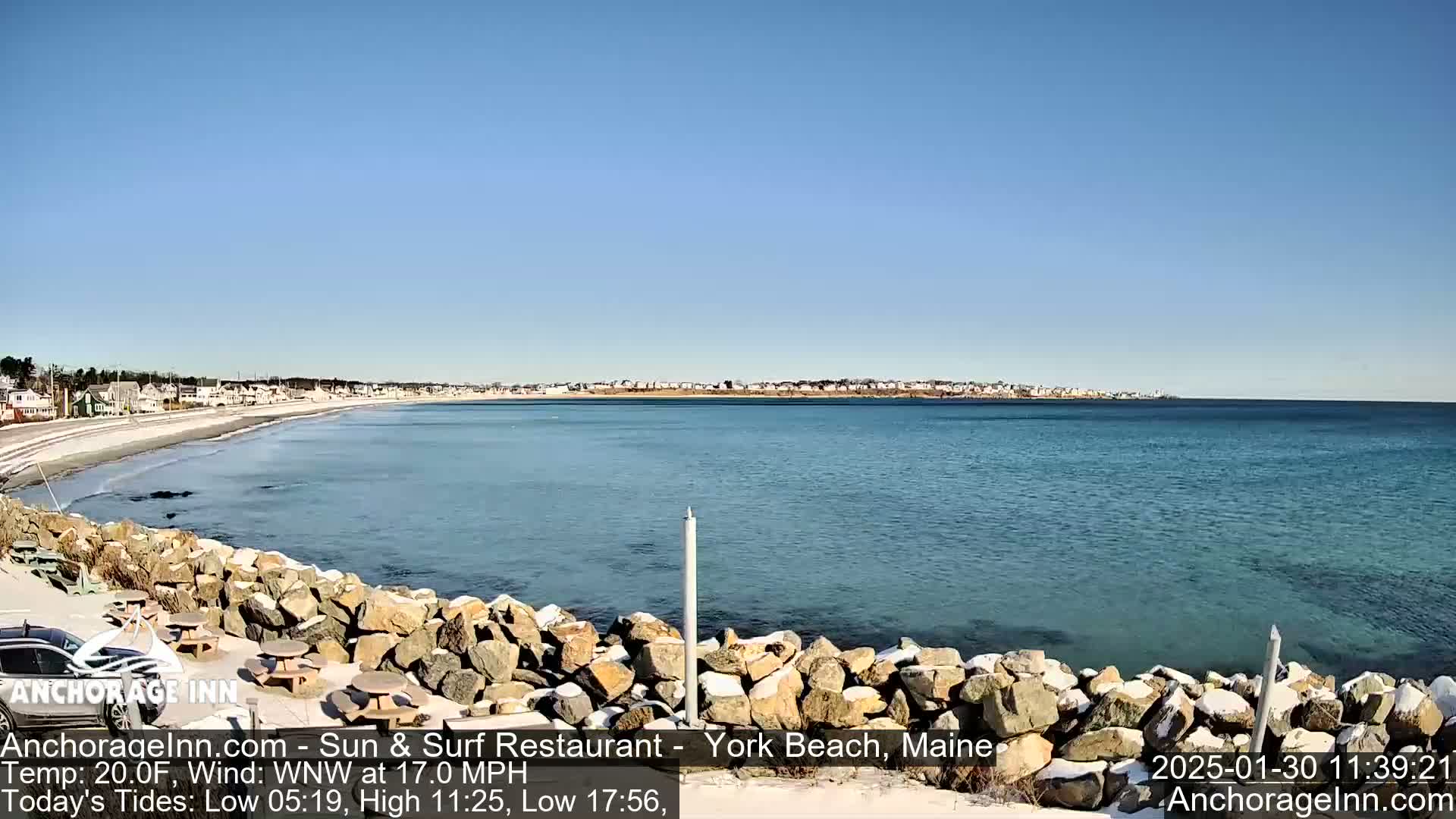 A tranquil ocean scene on a sunny day shows a sandy beach, calm water, and a rocky shoreline with some snow.