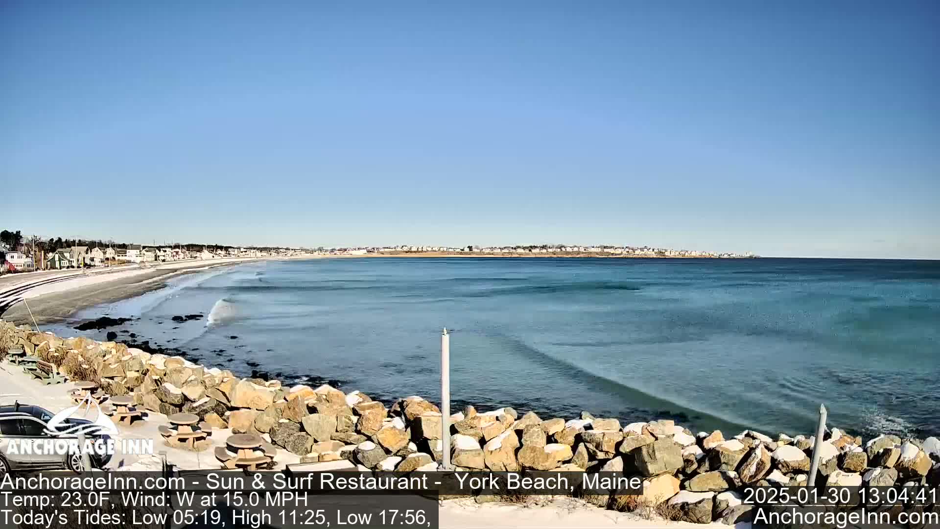 A partially snowy rocky shoreline overlooks a beach and ocean with small waves under a clear blue sky.
