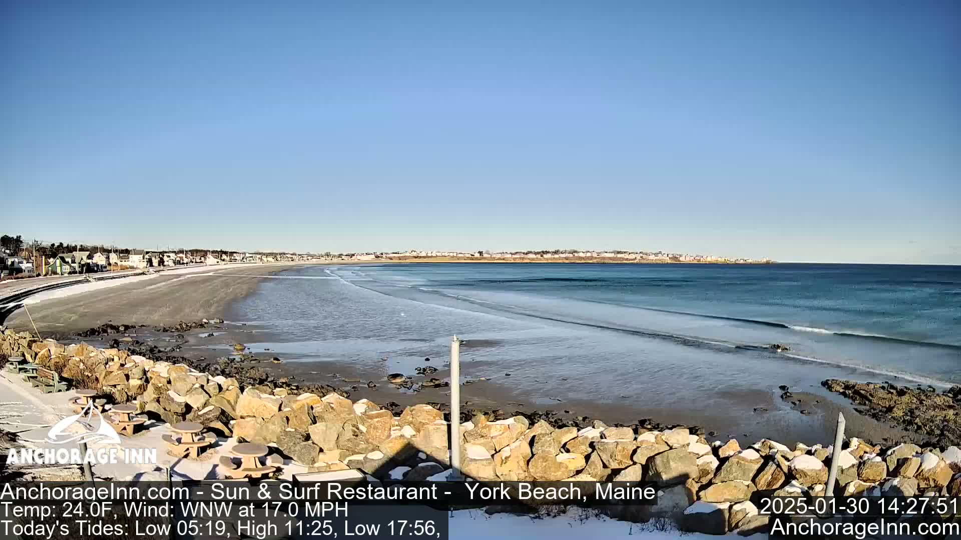 A sandy beach with gentle waves laps the shore under a clear blue sky, with some snow visible on the ground and rocks.